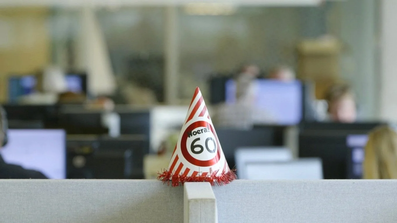 Feesthoed met "Hoera 60" in kantooromgeving, computers en mensen op de achtergrond.