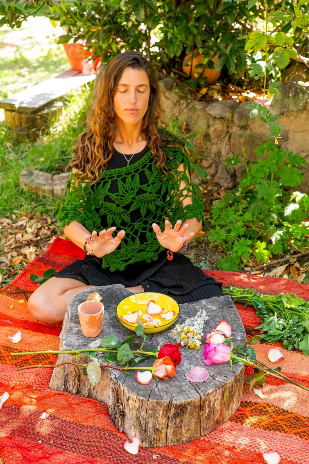 A woman sitting cross-legged outdoors on a colorful blanket, meditating with eyes closed, surrounded by flowers and greenery, with a small table holding a yellow bowl of flower petals, a cup, and some flowers in front of her.