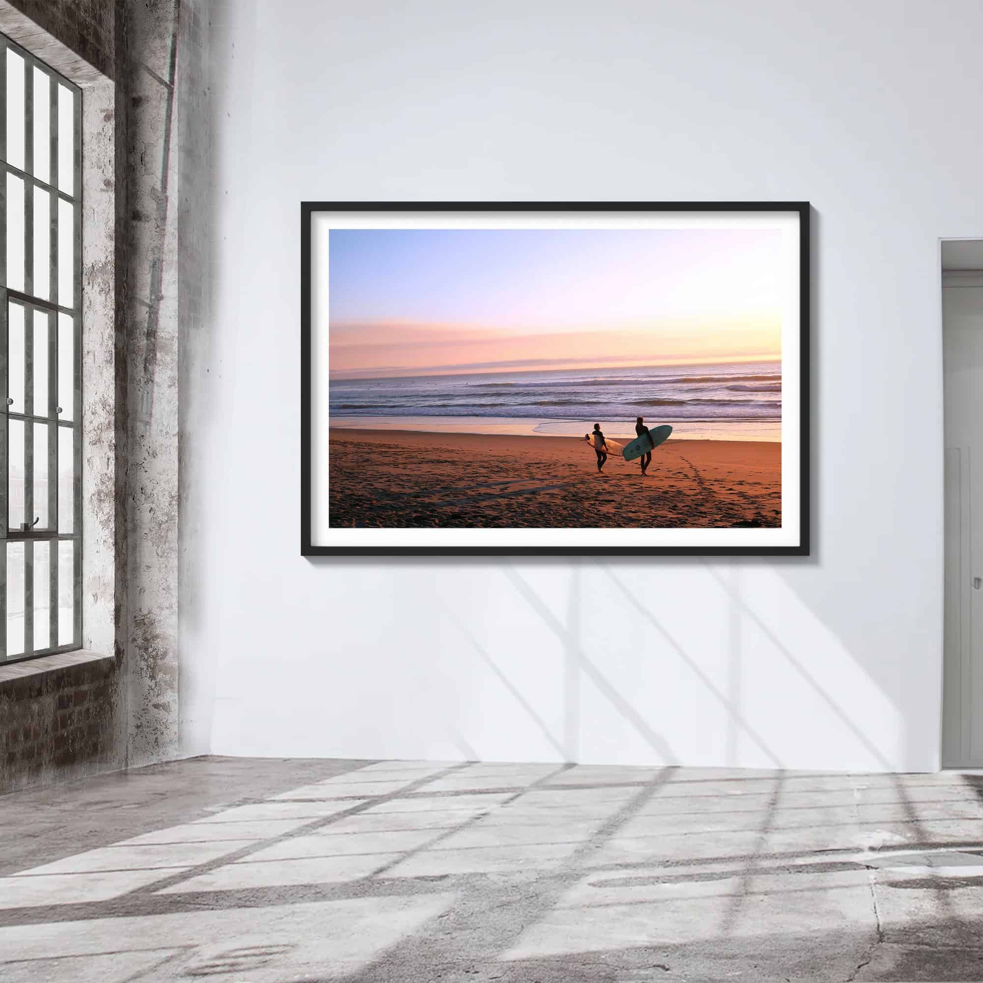 two surfers on the beach at sunset with pink, blue and yellow sky print with black wood frame on wall