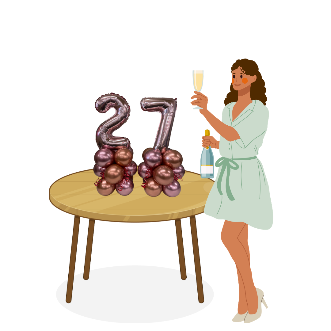 A woman in a white dress with a waist tie, holding a champagne glass and a bottle, standing next to a wooden table decorated with pink and metallic balloons shaped as the number 27, indicating a celebration or birthday party.