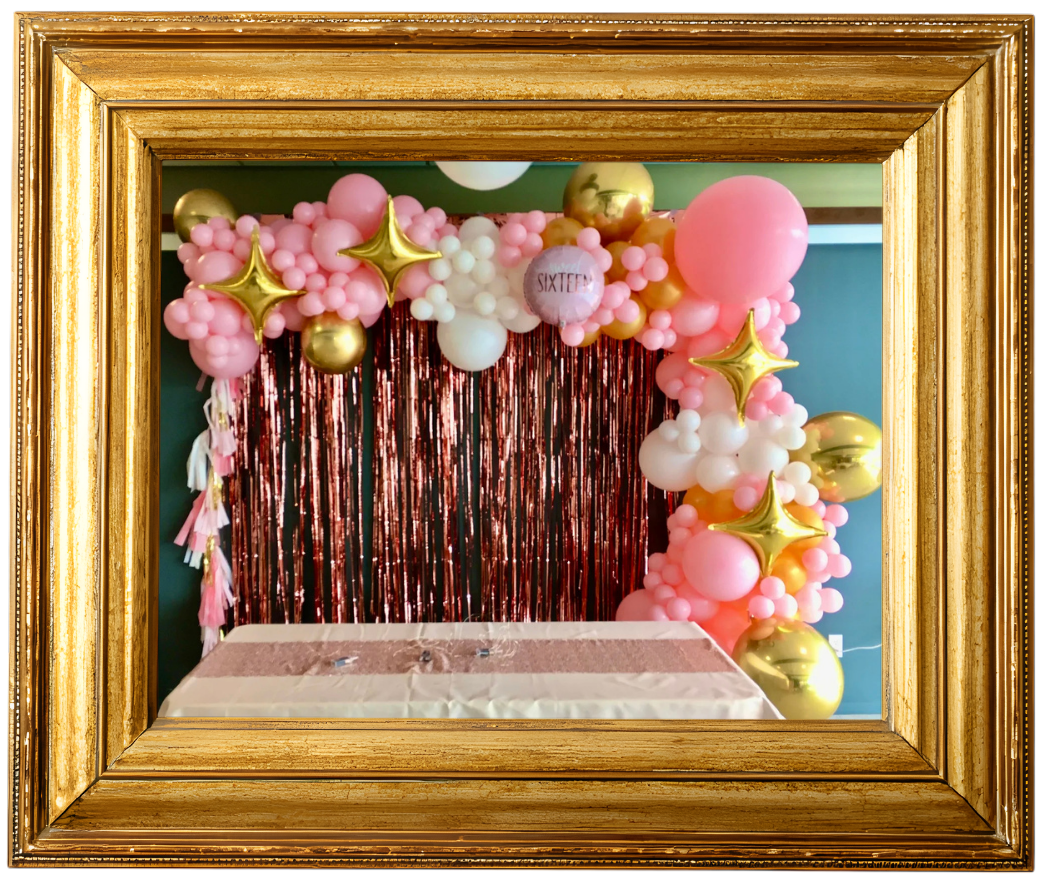 Decorative frame surrounding a birthday party backdrop with pink, white, and gold balloons, pink metallic fringe curtain, and a sign that reads 'SIXTEEN'