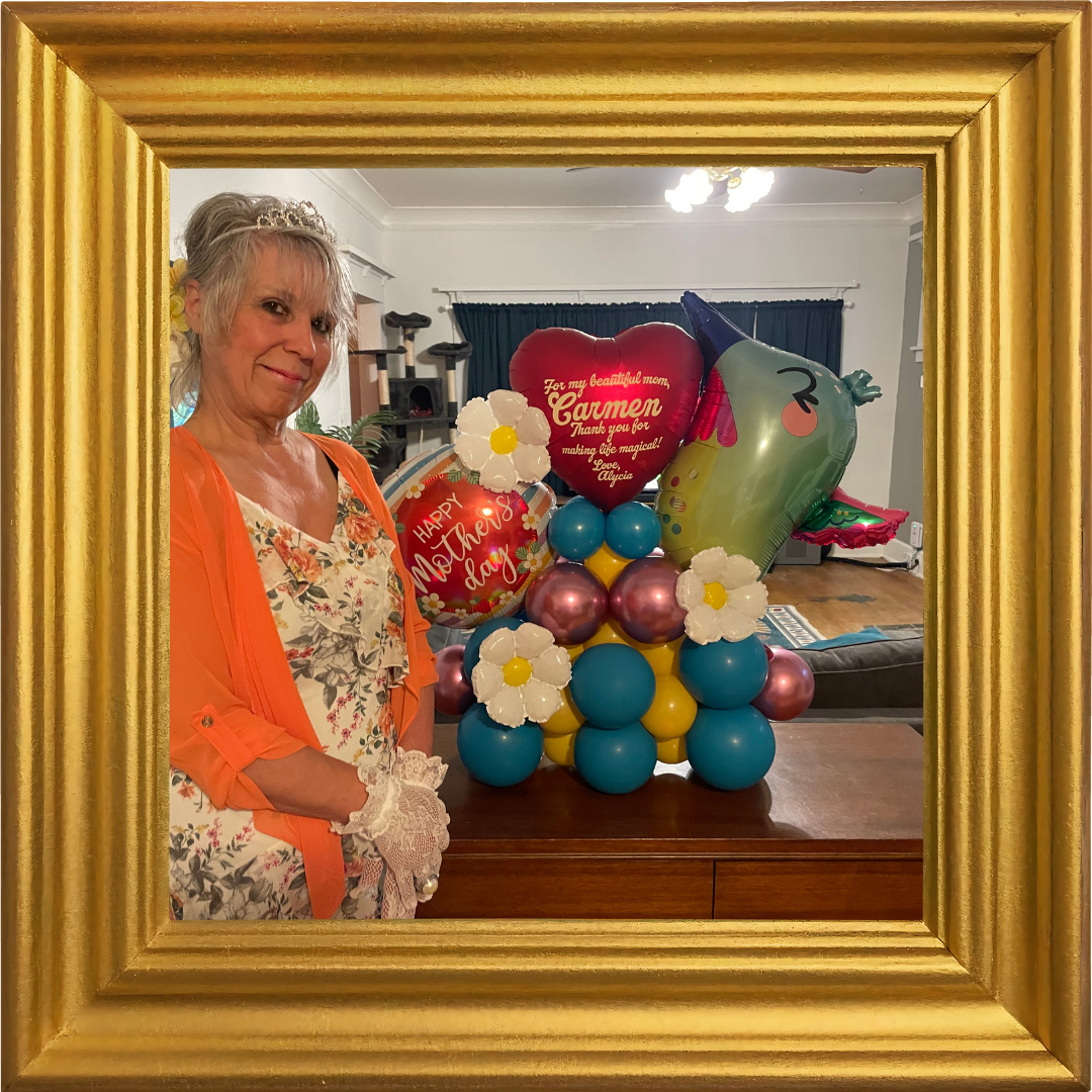 A woman dressed in floral and orange, wearing a small tiara and lace gloves, stands beside a table decorated with colorful balloons and flowers. The balloons include one with a happy mother's day message, a heart-shaped balloon with a message for Carmen, and a unicorn-shaped balloon. The scene appears to be in a living room with a sofa, curtains, and a cat tree in the background, viewed through a gold picture frame.