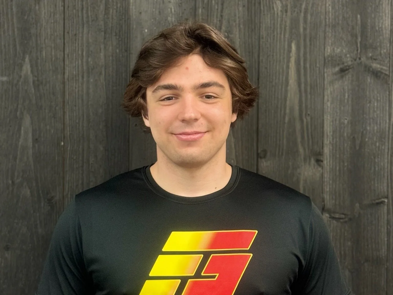 Man wearing a black shirt with red and yellow logo, standing against a wooden wall background.