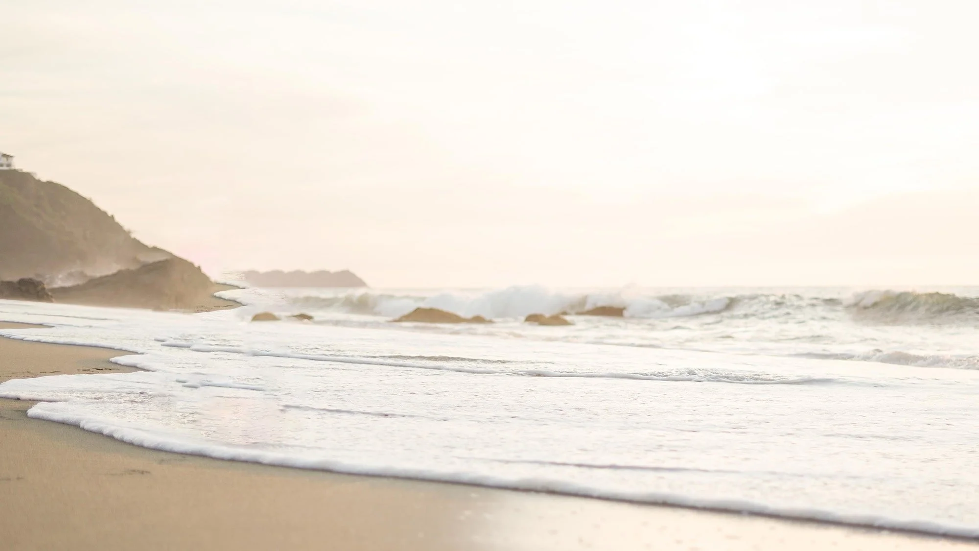 A peaceful beach scene with gentle waves washing onto the sandy shore, cliffs in the background, and a bright, slightly hazy sky.