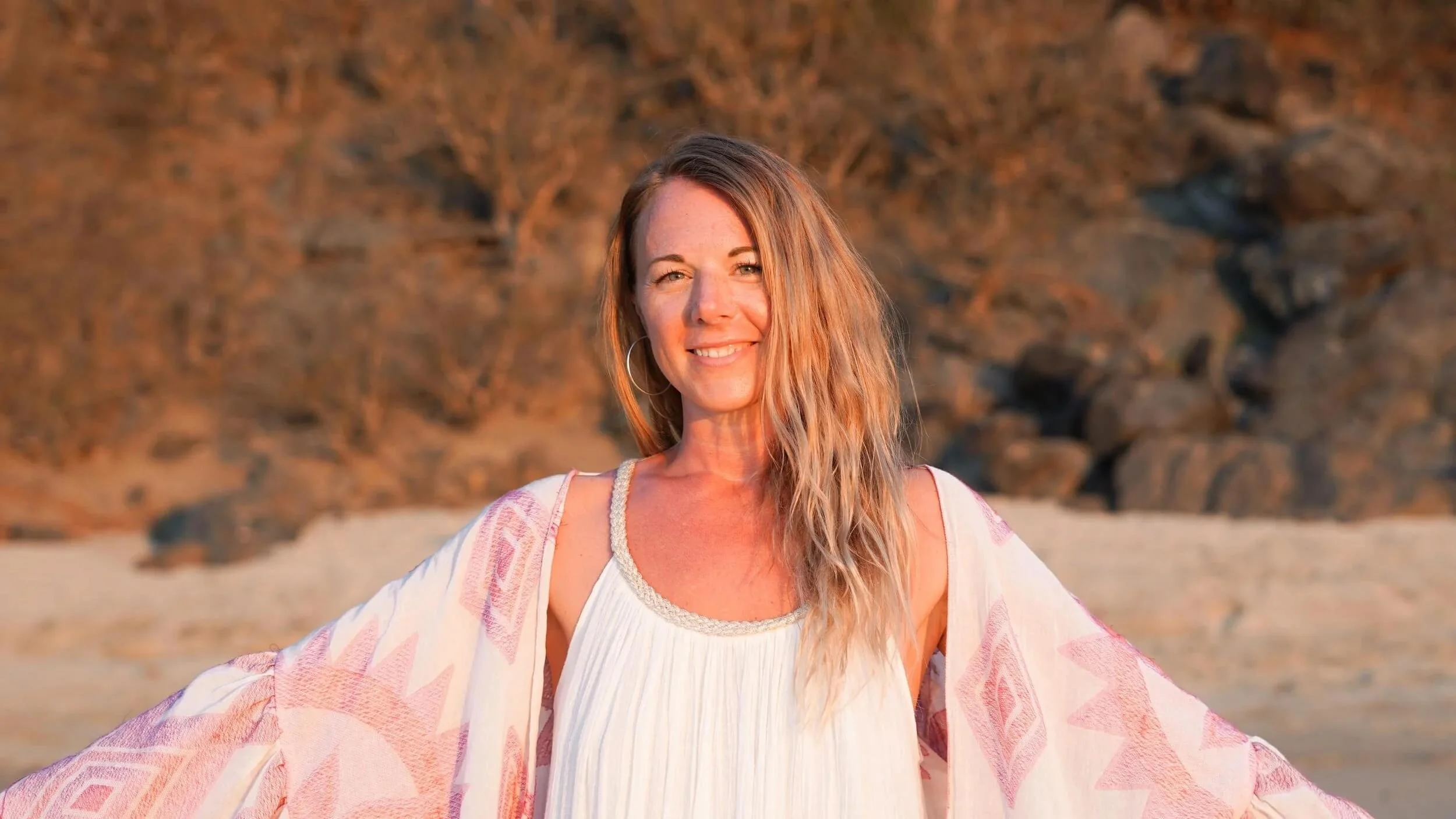 A woman with long, wavy hair wearing a white dress and a pink shawl standing on a beach during sunset.