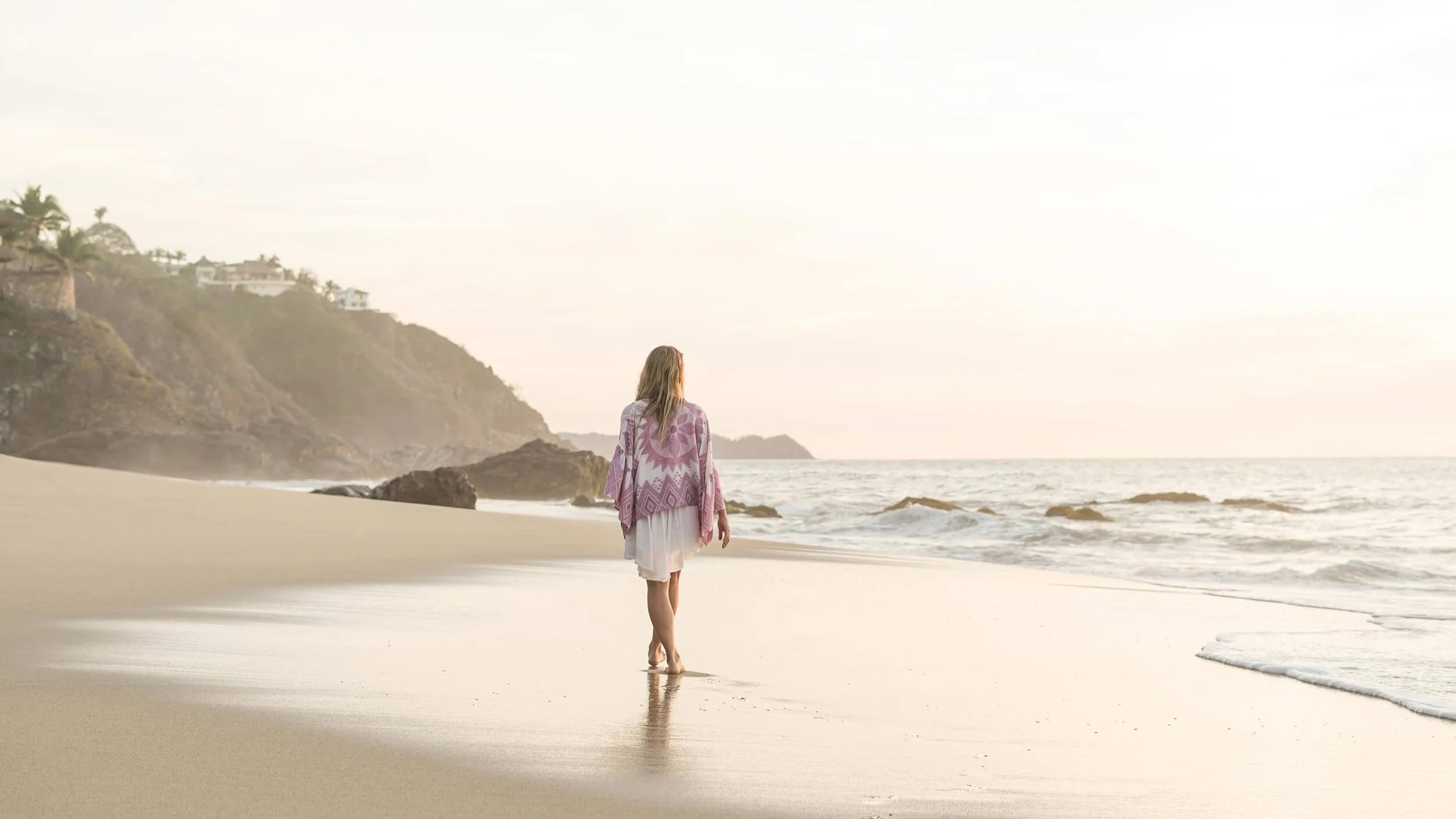 A woman in a pink and white patterned shawl and white dress walking on a beach at sunset, with cliffs and houses in the background.