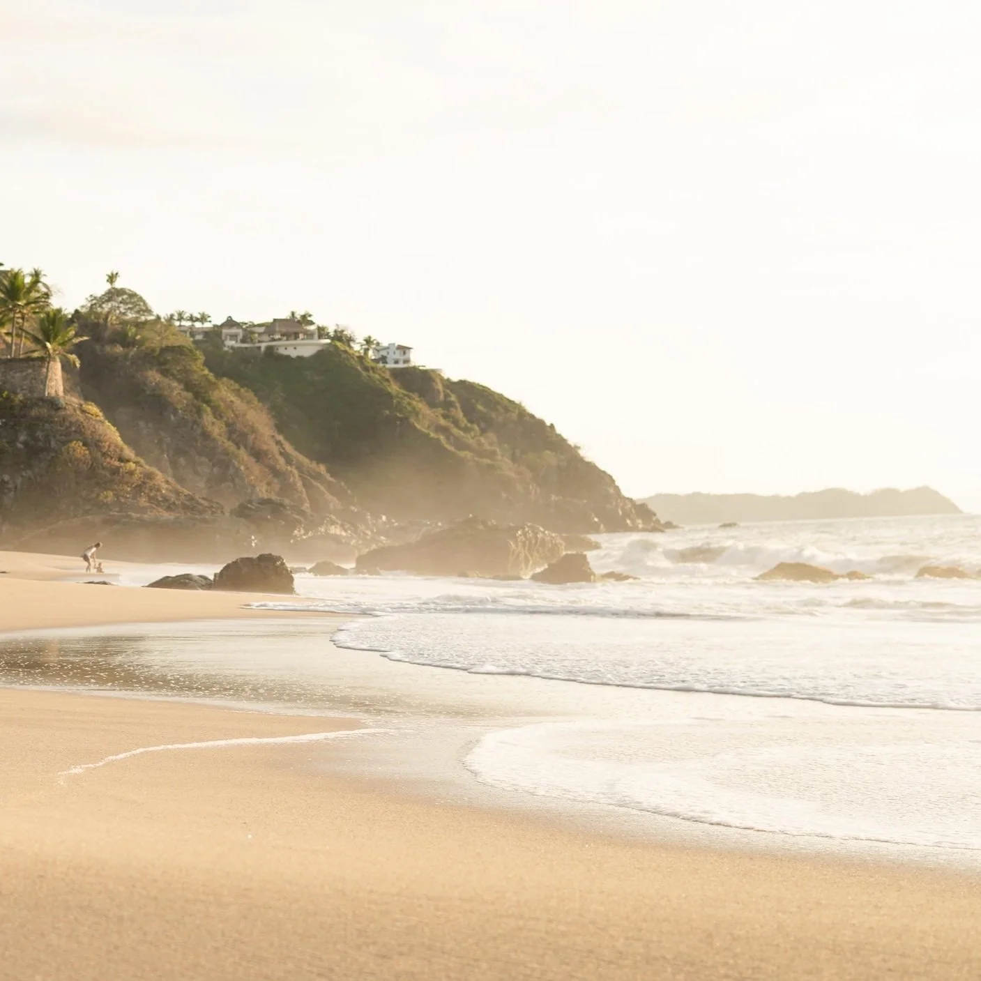 A sandy beach with rocks, waves, and a person crouching near the water, overlooking a hillside with houses and palm trees in the distance.