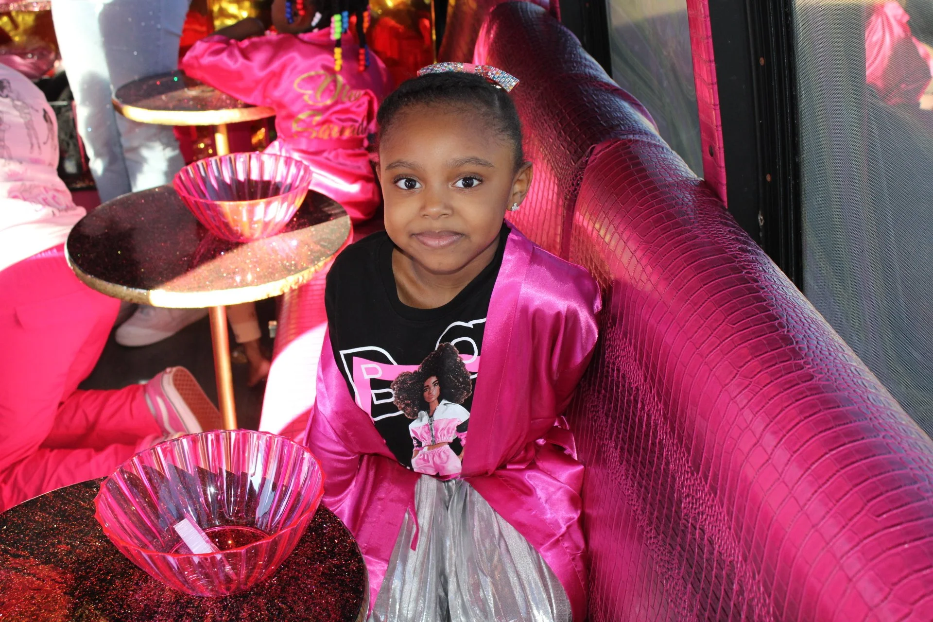 A young girl sitting at a table in a pink-themed party venue, wearing a black t-shirt with a graphic of a woman and the word 'Barbie,' a pink satin jacket, and a shiny silver skirt. The table has pink glass bowls with glittery black surfaces.