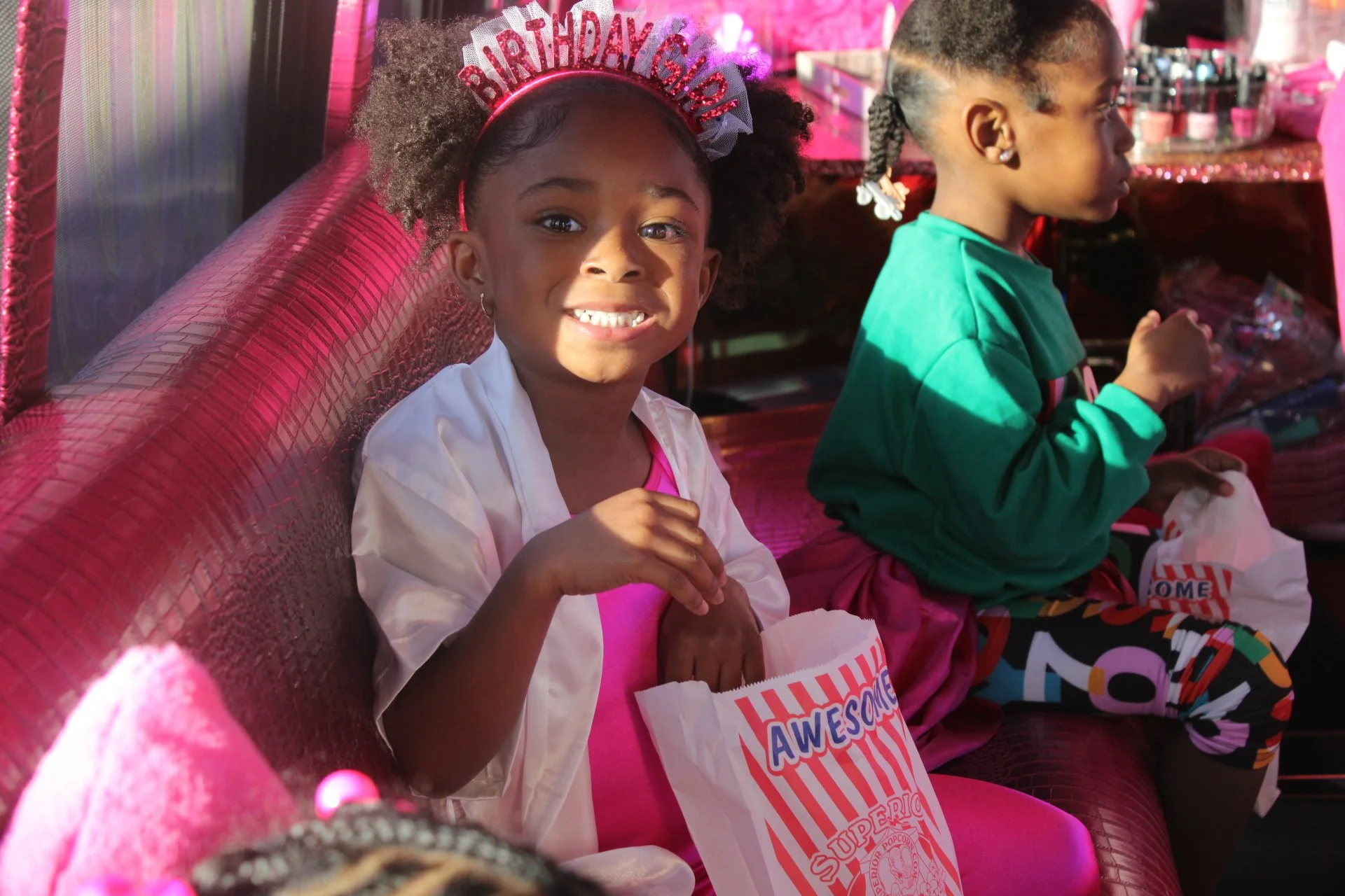 A young girl wearing a birthday crown labeled 'Birthday Girl' and a pink dress, smiling and holding a popcorn bag at a birthday party with pink decorations, sitting on a pink bench.