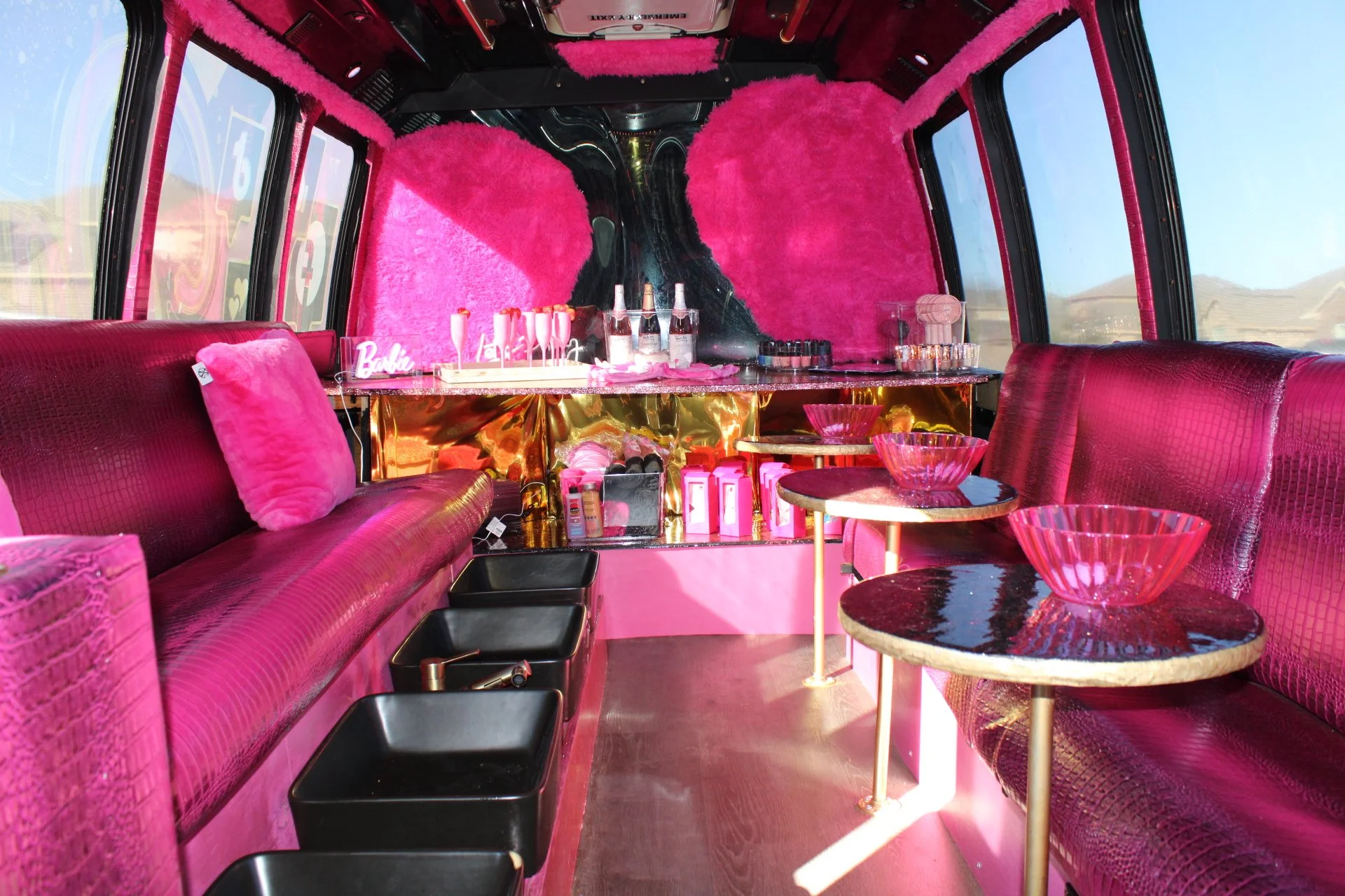 Inside of a pink-themed party bus with velvet and faux fur decor, pink couches, small tables, and bottles of champagne, with a Barbie sign on the counter.