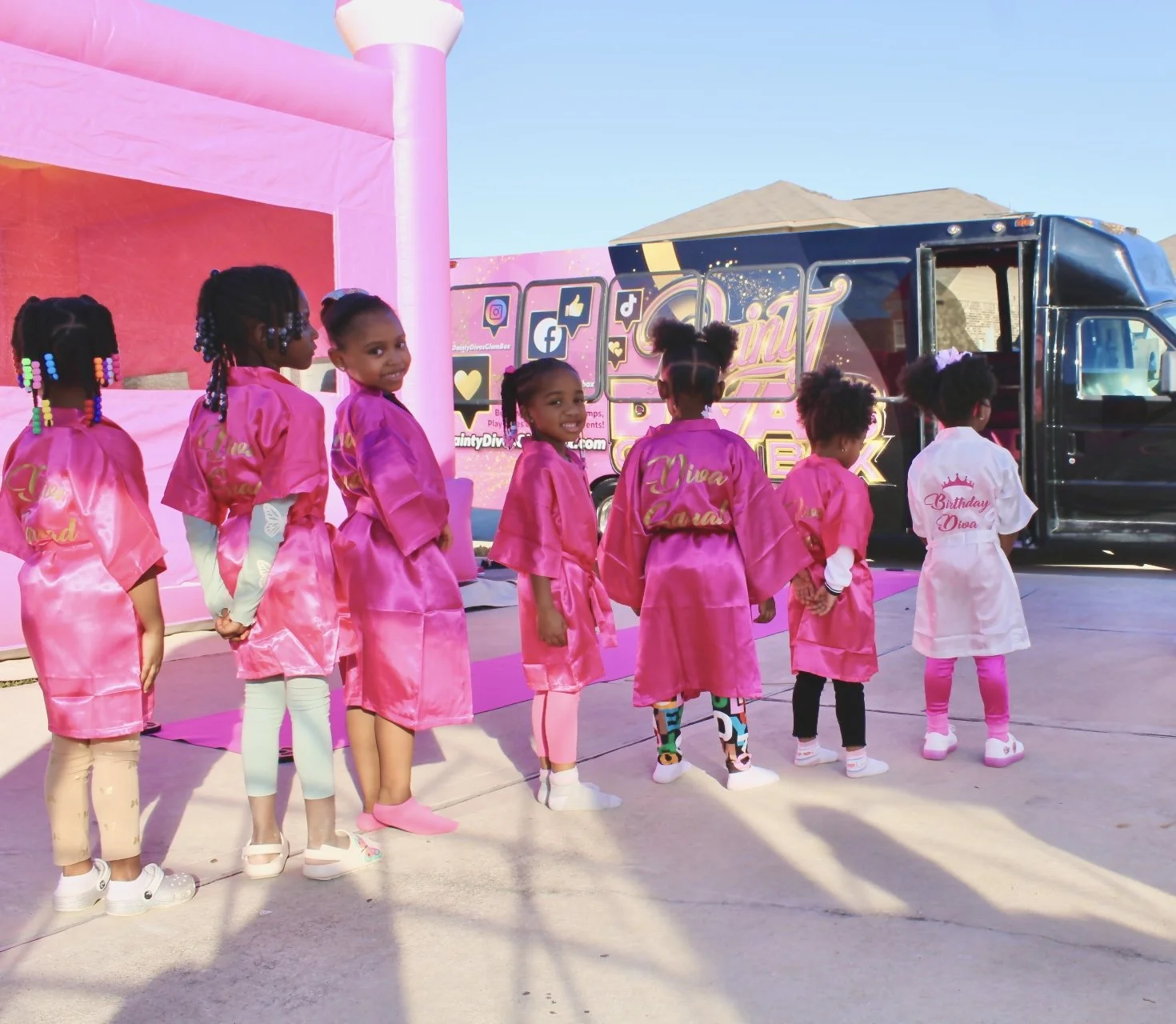 Children dressed in pink satin robes standing in line outdoors during a celebration, with a food truck and pink decorations in the background.