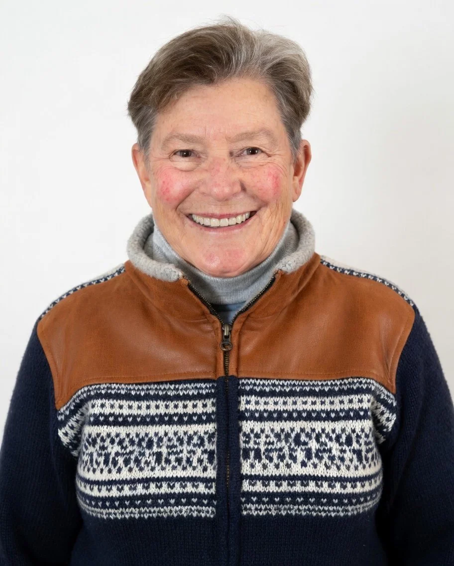 Smiling elderly woman with short gray hair, wearing a gray turtleneck, a brown leather and knit sweater, and standing against a white background.