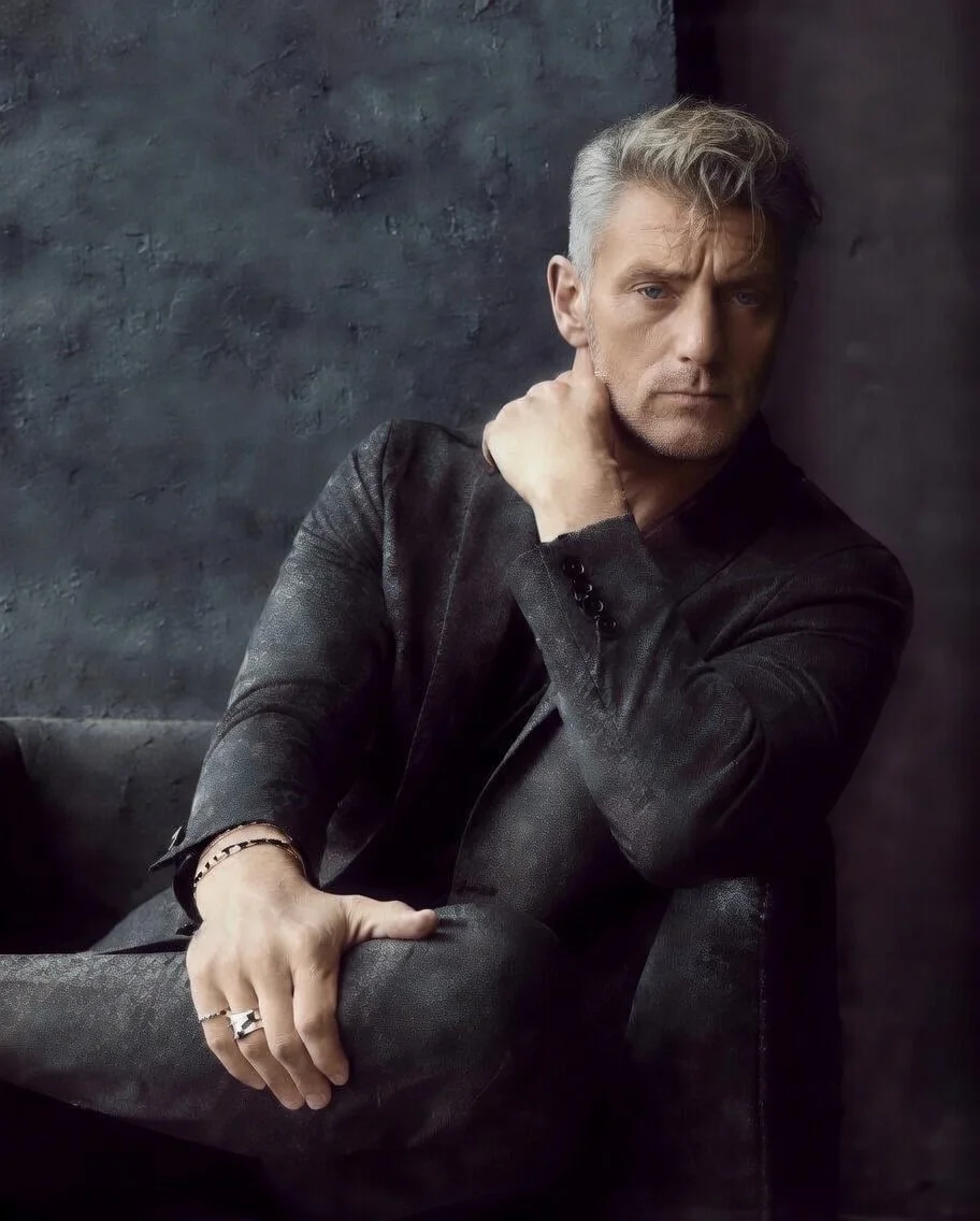 A middle-aged man with gray hair sitting against a dark wall, wearing a dark blazer, with his left hand resting on his knee and his right hand touching his neck, looking serious.