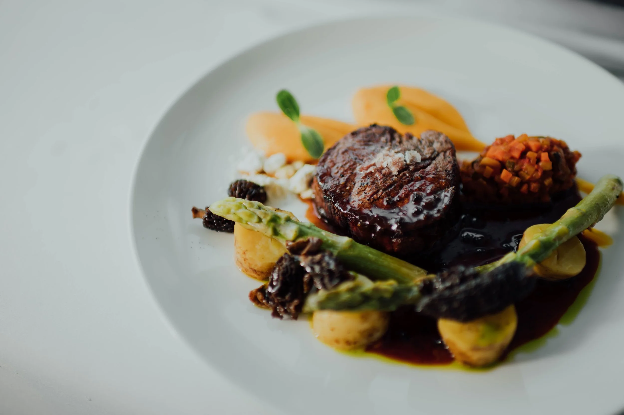 Gourmet plated dish features a grilled steak topped with sauce, accompanied by asparagus, yellow puree, carrot and vegetable croquette, and garnished with small green leaves.