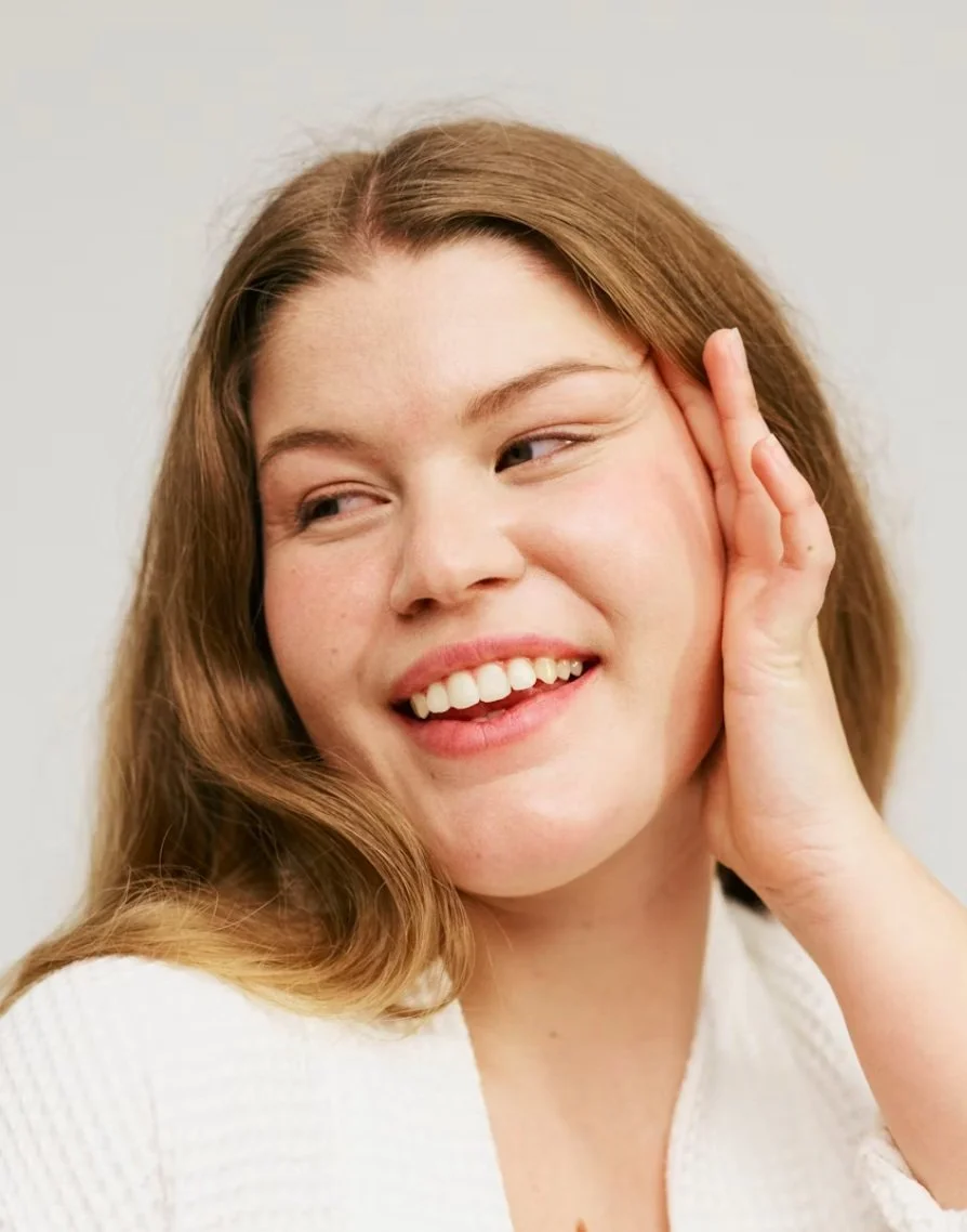 A woman touching her ear and smiling with her teeth visible, with long wavy hair and a light-colored top.