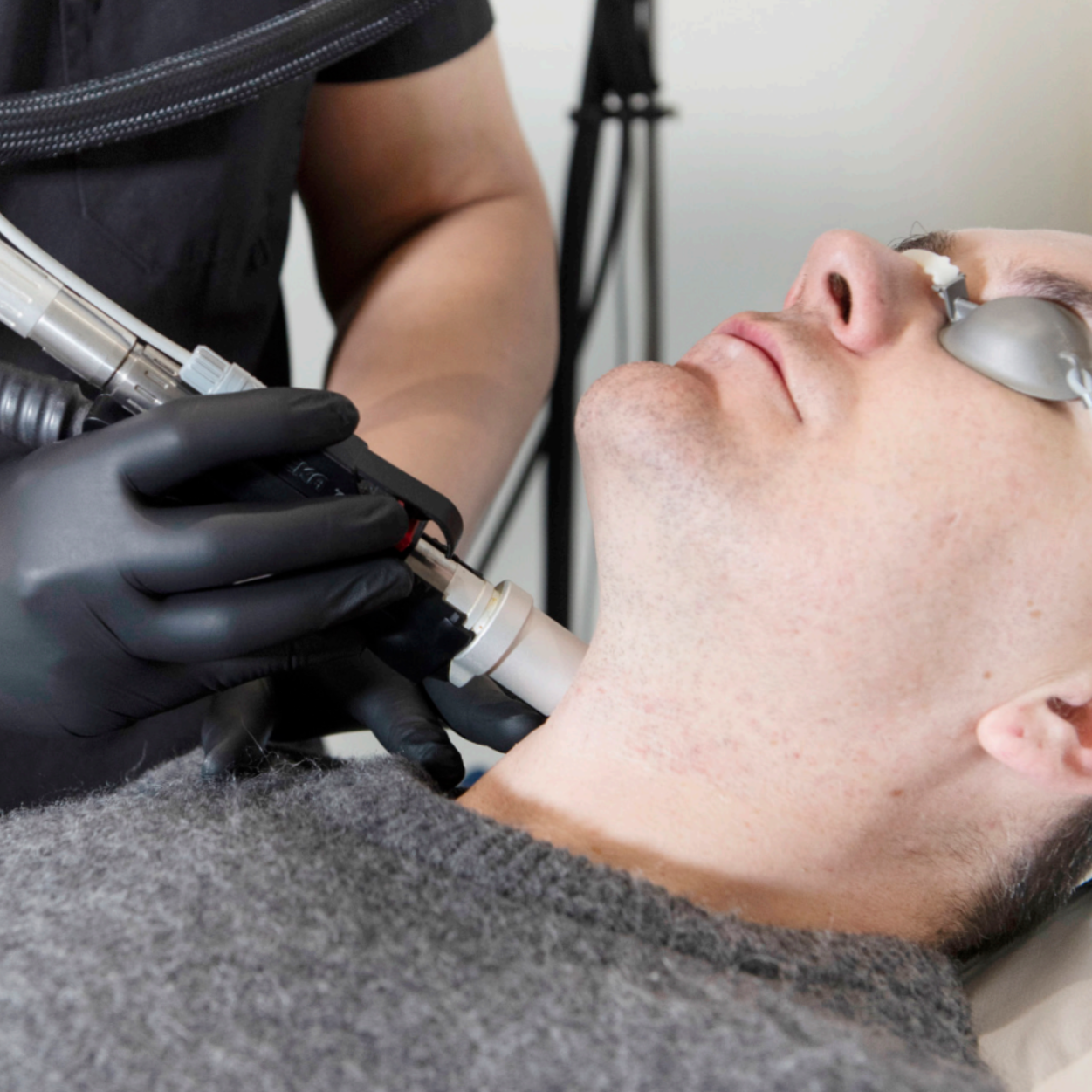 A man receiving a cosmetic or dermatological treatment on his neck using a specialized device held by a medical professional wearing black gloves.