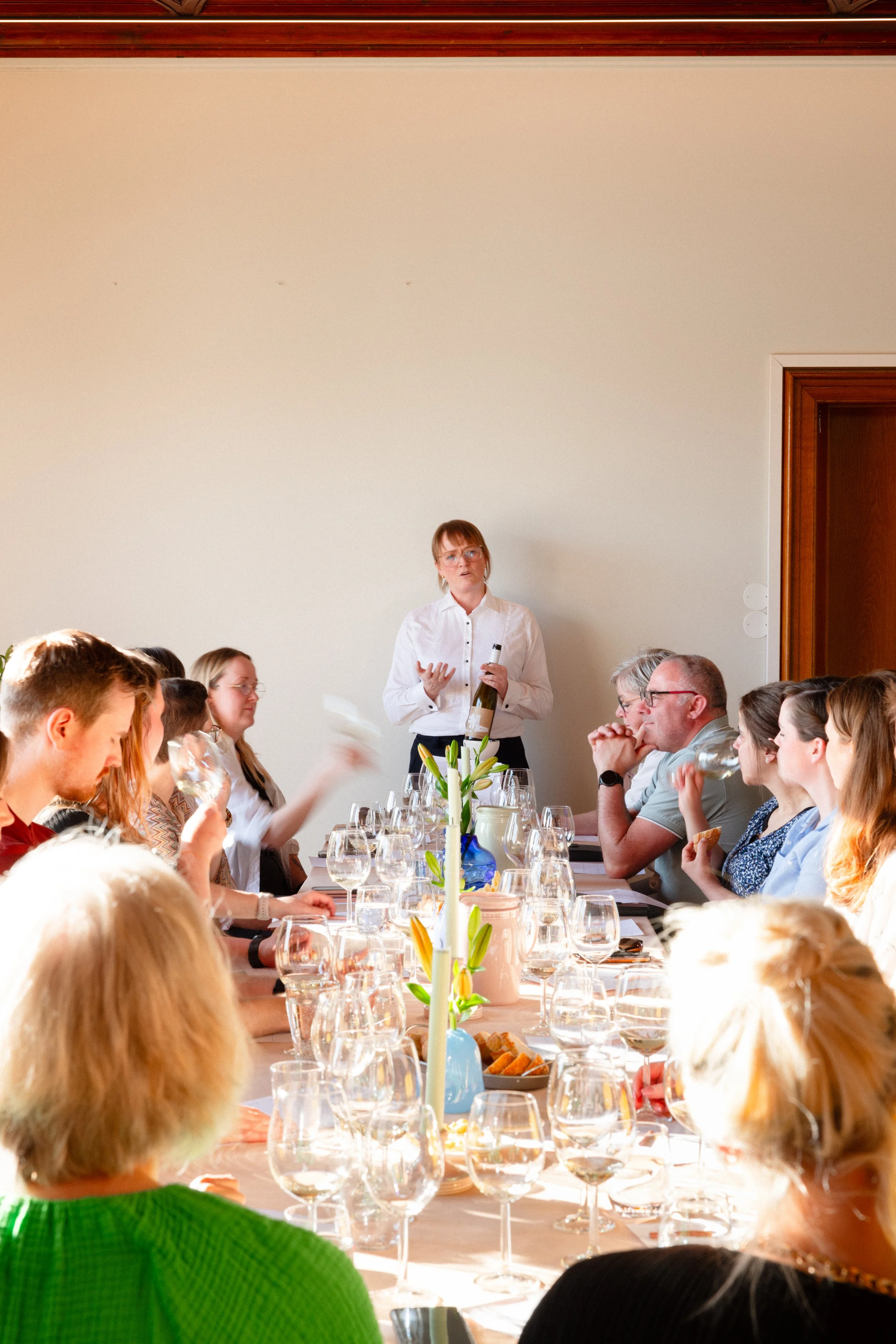 En gruppe mennesker sitter rundt et langbord og deltar i en vin- eller smaksprøvetilstelning, mens en kvinne står foran og snakker.