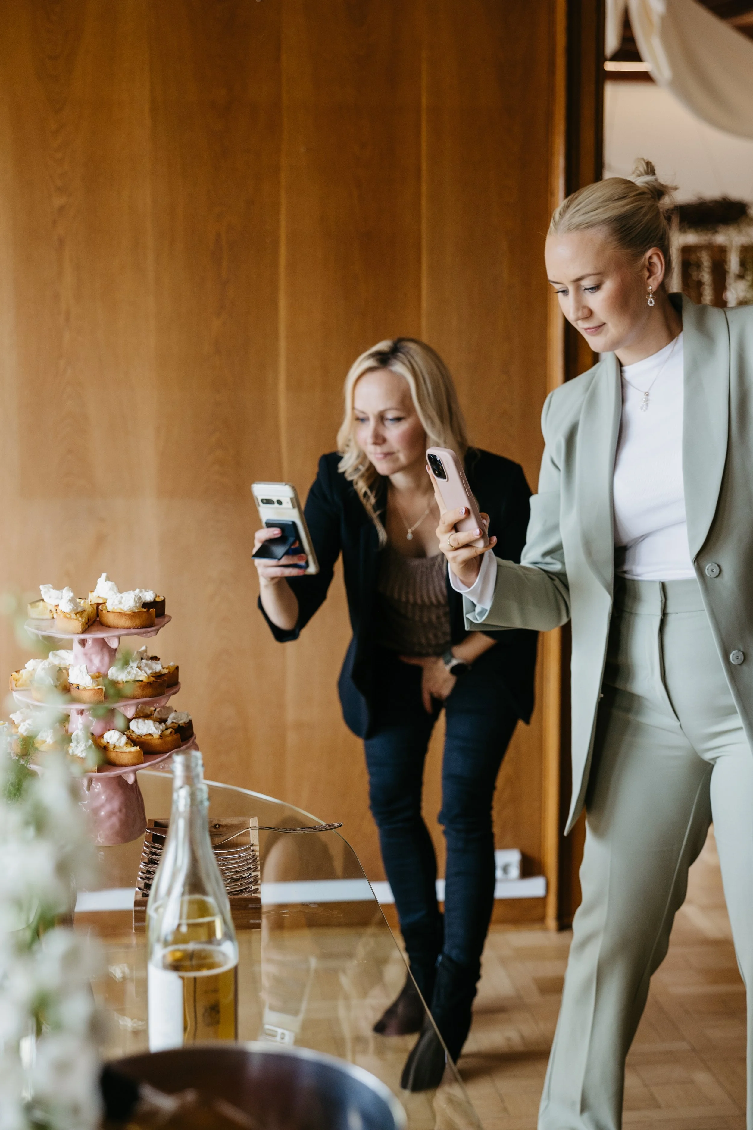 To kvinner tar bilder av en kake med små cupcakes på en 3-etasjers kakefat under en sammenkomst.