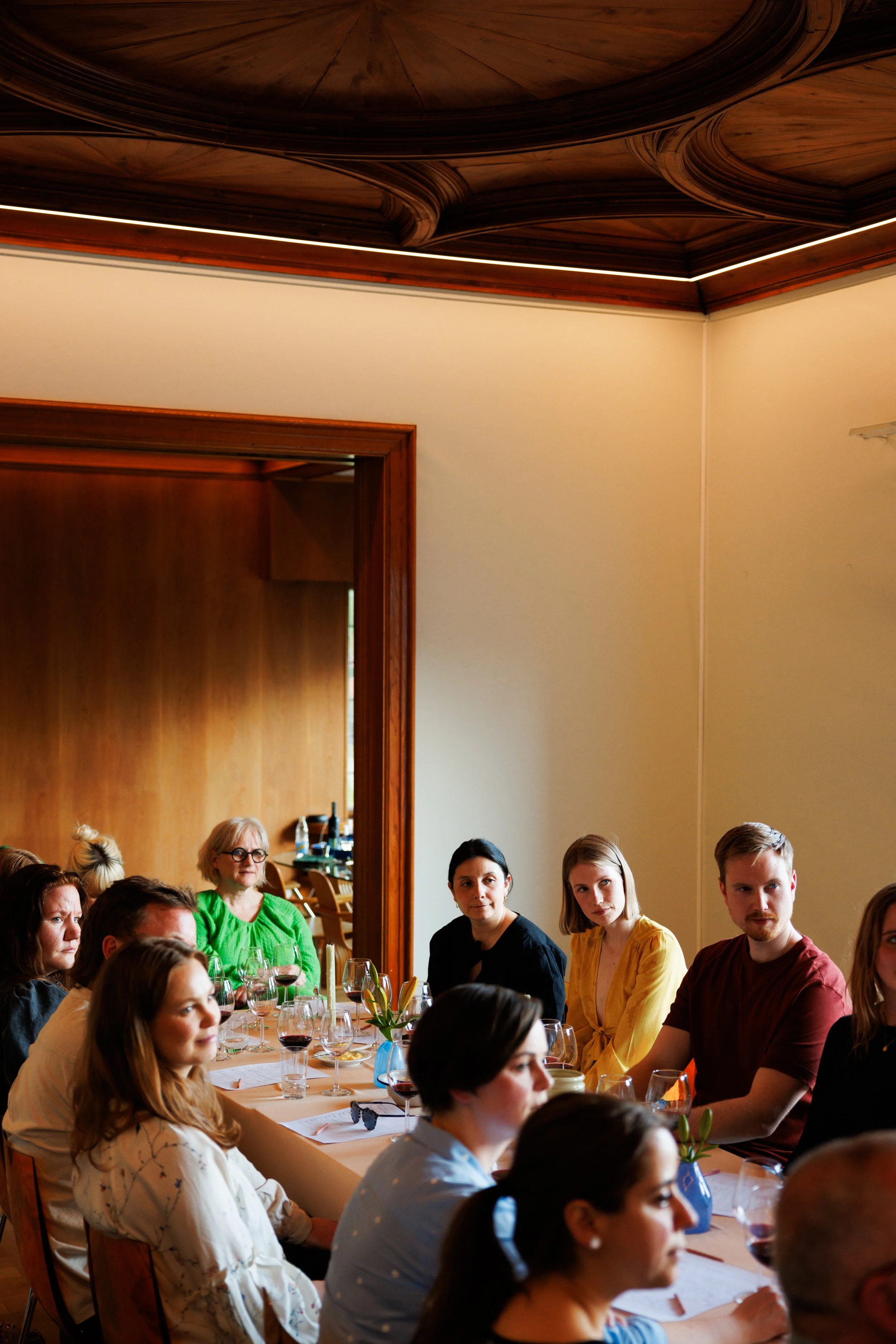 Personer som sitter rundt et langbord under et møte eller en sammenkomst i et rom med trepanel og lys vegg.