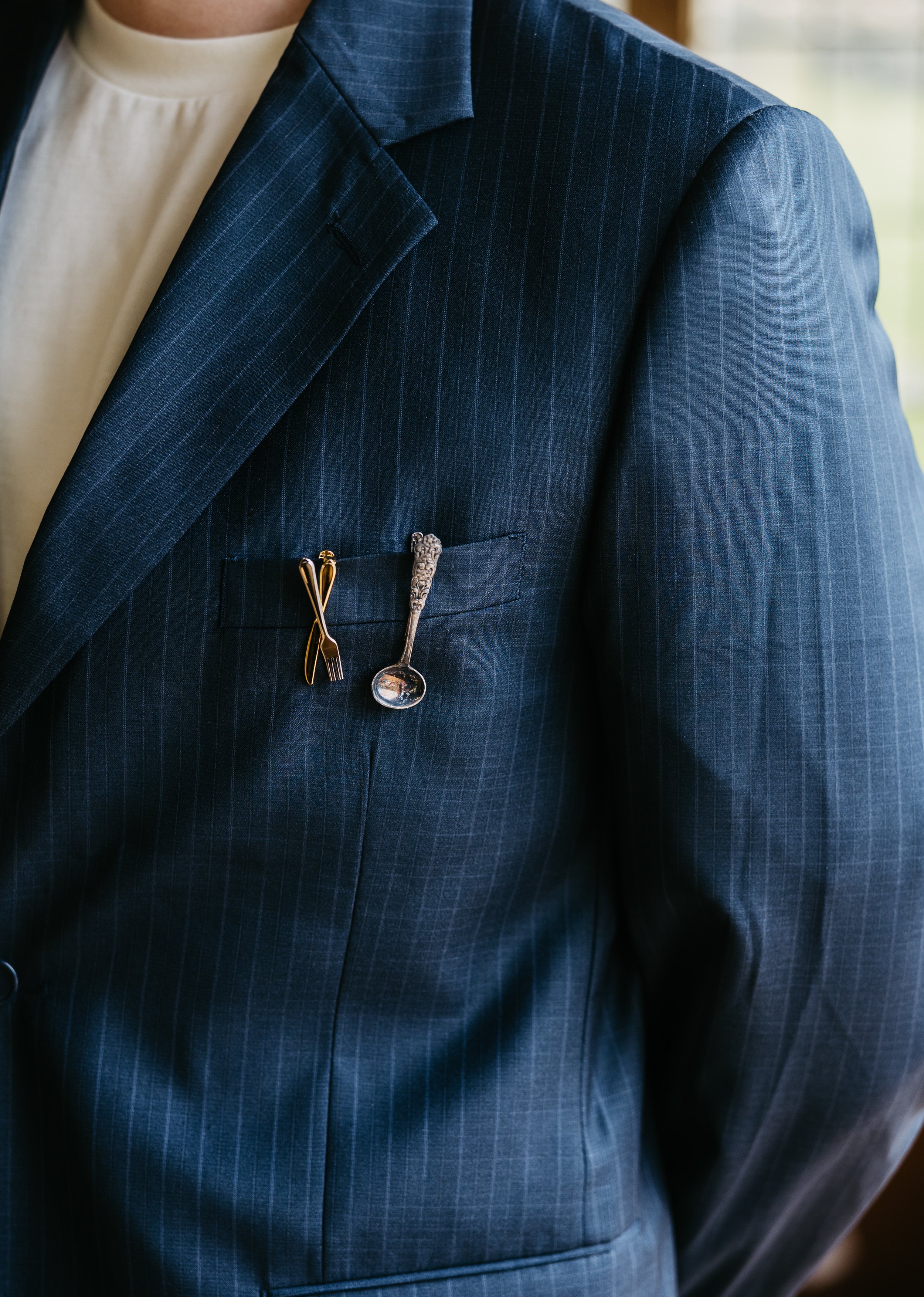 Nærbilde av en dressjakke med to små figurer av bestikk festet i brystlommen, en liten gullfarget gaffel og en sølvskje.