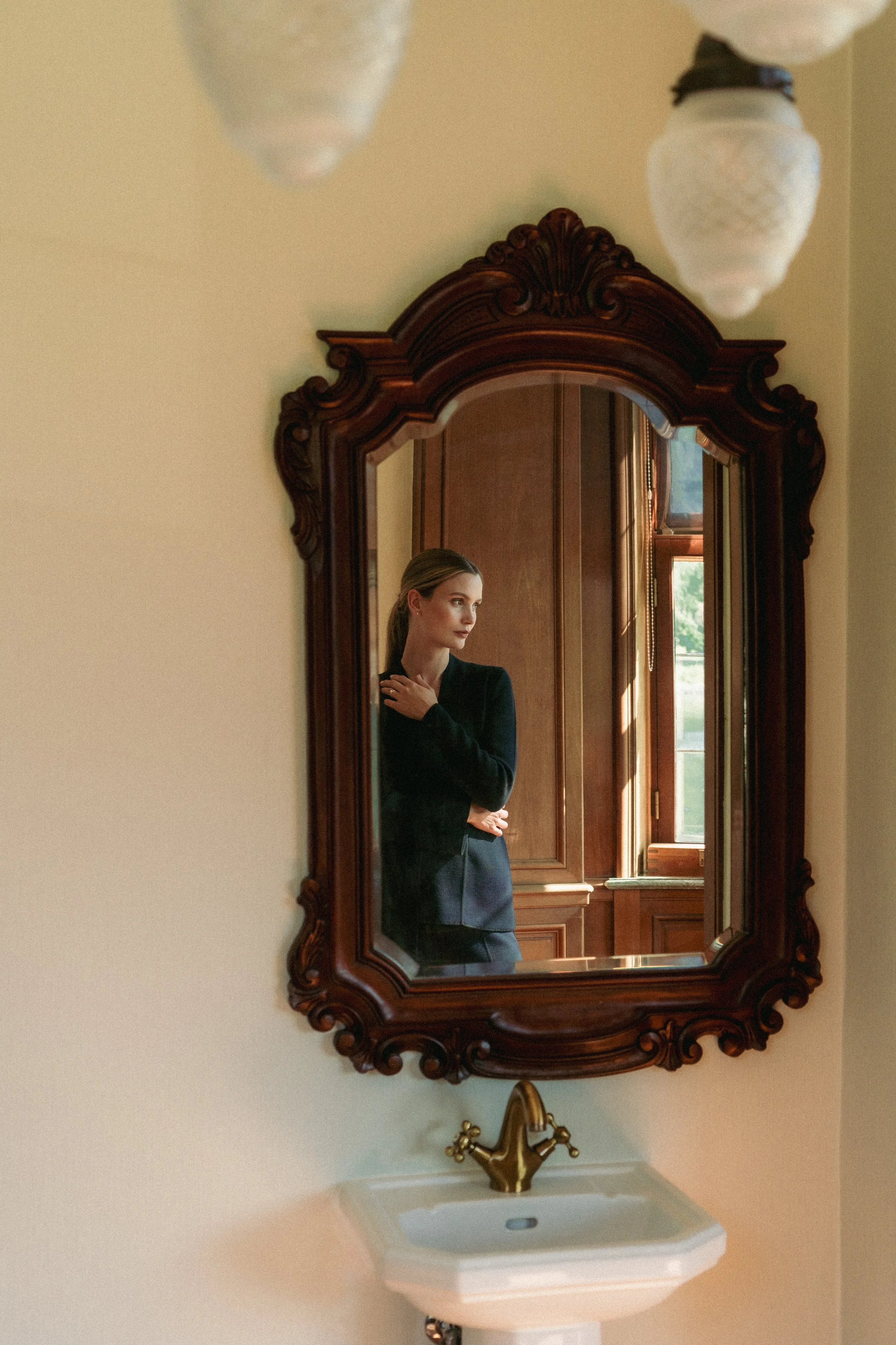 Et speil som reflekterer en kvinne som står i et rom med trevinduer, og sitter over en hvelvet vask med gullhåndtak.