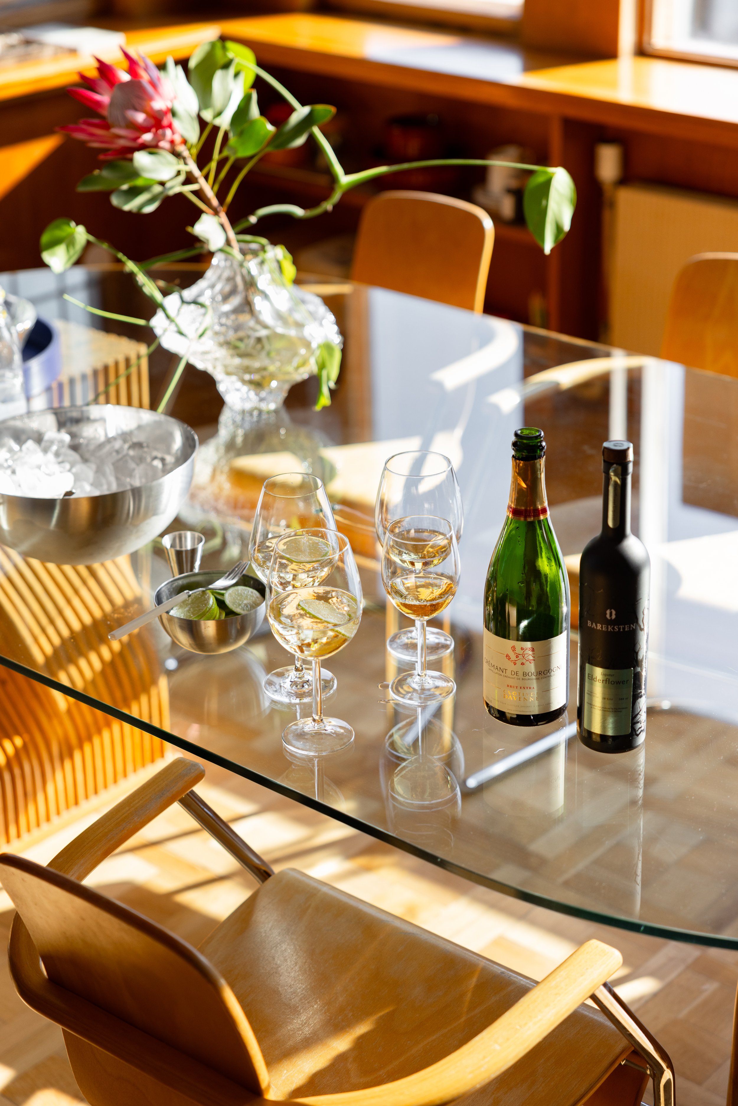 En glassadvask på et spisebord med flere tomme vin- og musserende vinstyper, en vinflaske, og en stor vase med en blomsterbukett. Sollys skinner inn gjennom et vindu.