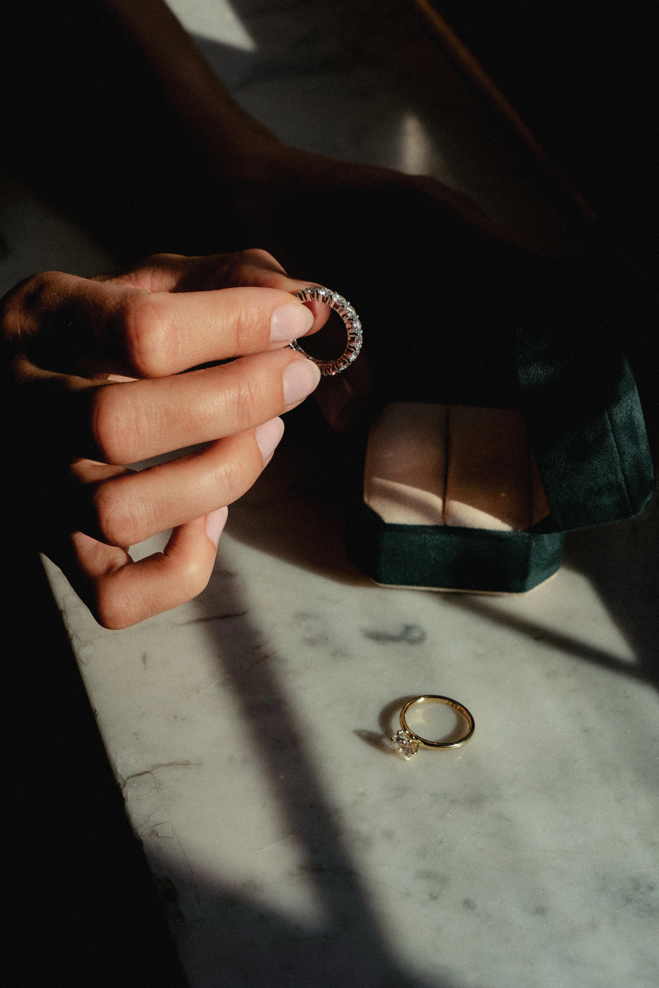 Hånd holder en ring med diamanter, med en boks med mer smykker og en enkelt gullring på et Marmorbord.