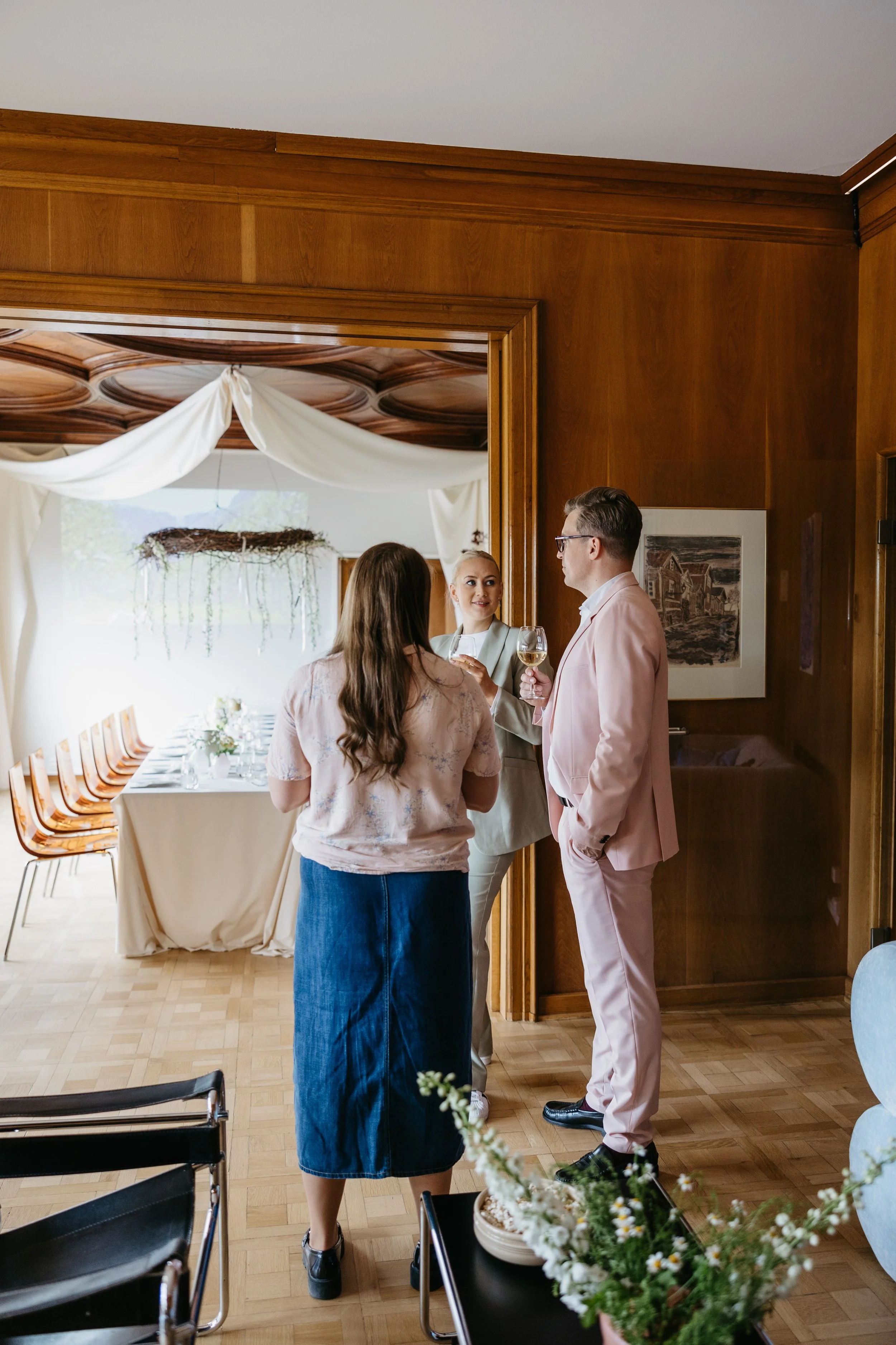 Tre personer som snakker og smiler i et rom med tre kvister i en rund vase i forgrunnen.