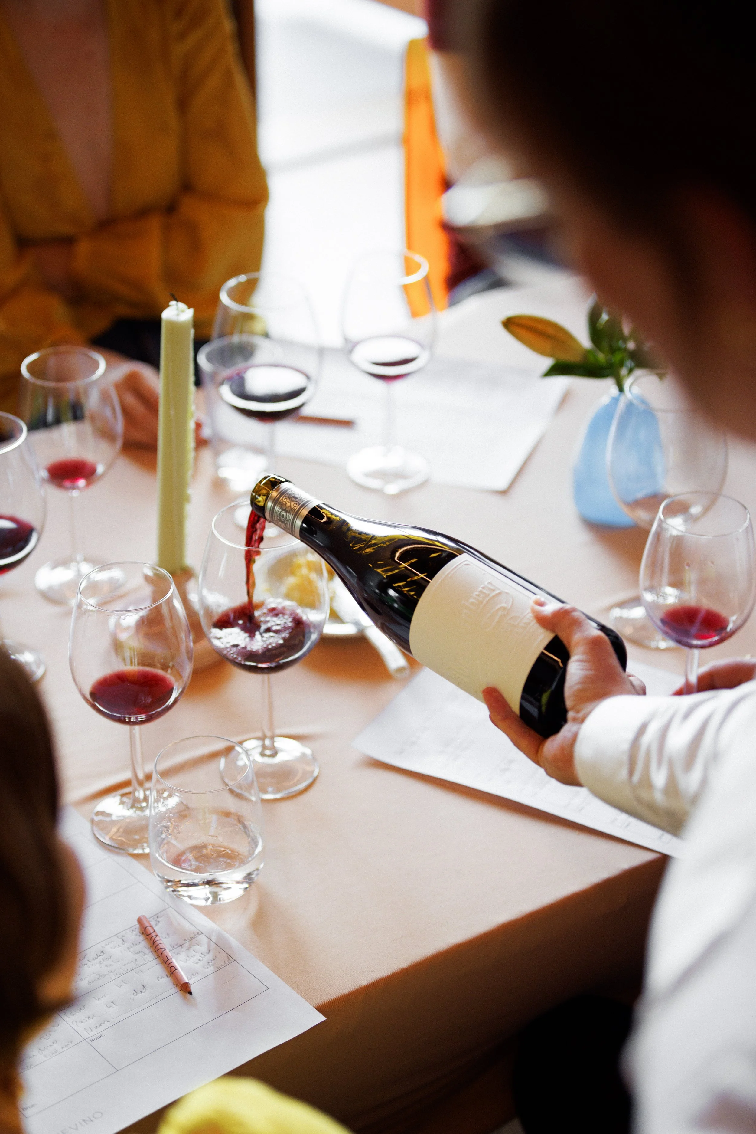 En person heller rødvin i glass, omgitt av flere andre glass med rødvin på et bord, med en lys grønn lysestake og en blomst i en blå vase.