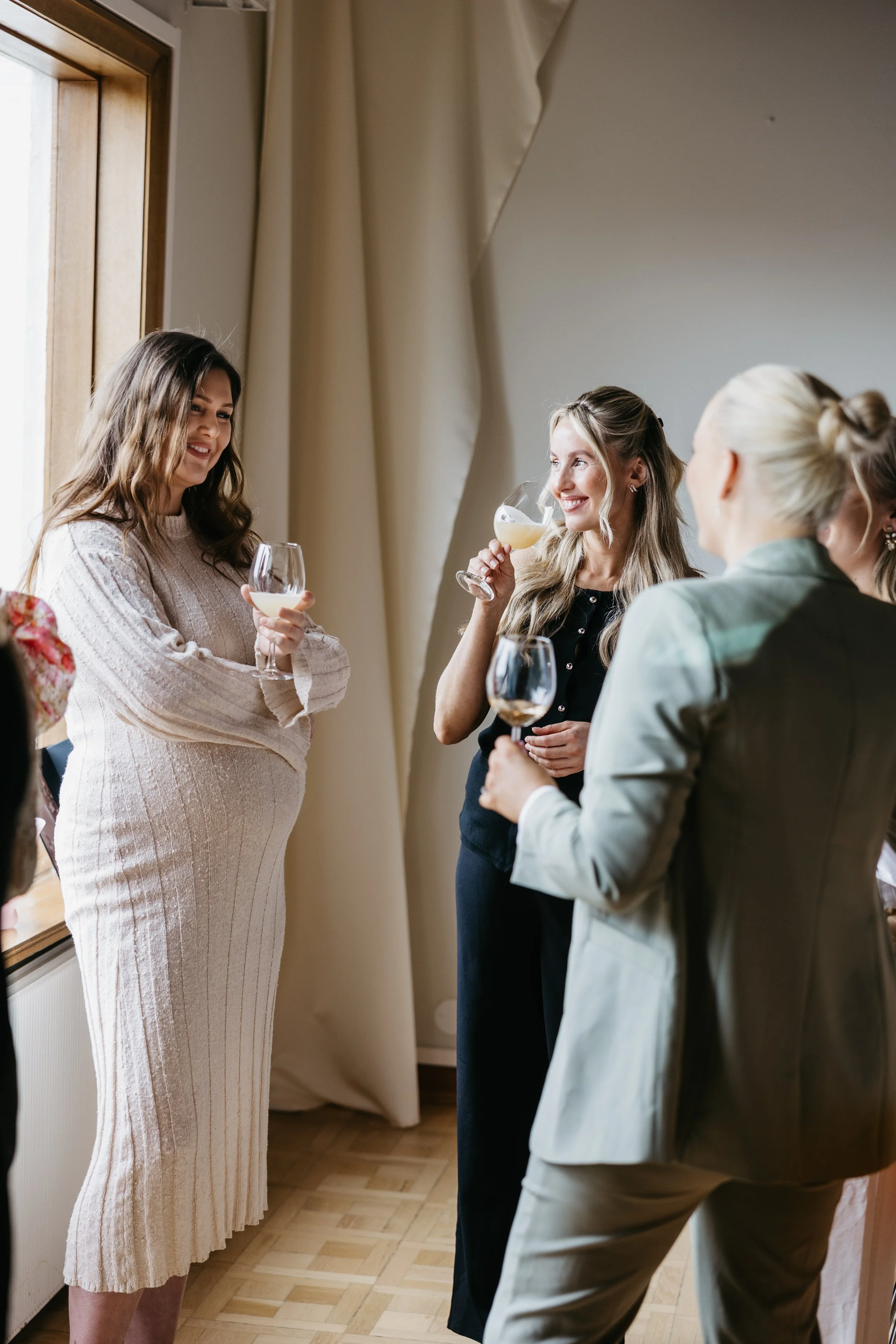 Kvinner i festantrekk som drikker vin og snakker innendørs, med klesskap og vindu i bakgrunnen.