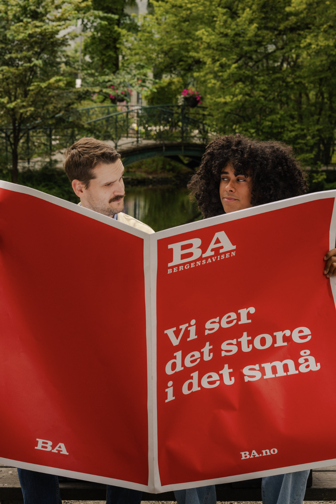 To mennesker holder et rødt skilt med teksten "Vi ser det store i det små" foran en park med grønne trær og en liten bro over en liten elv.