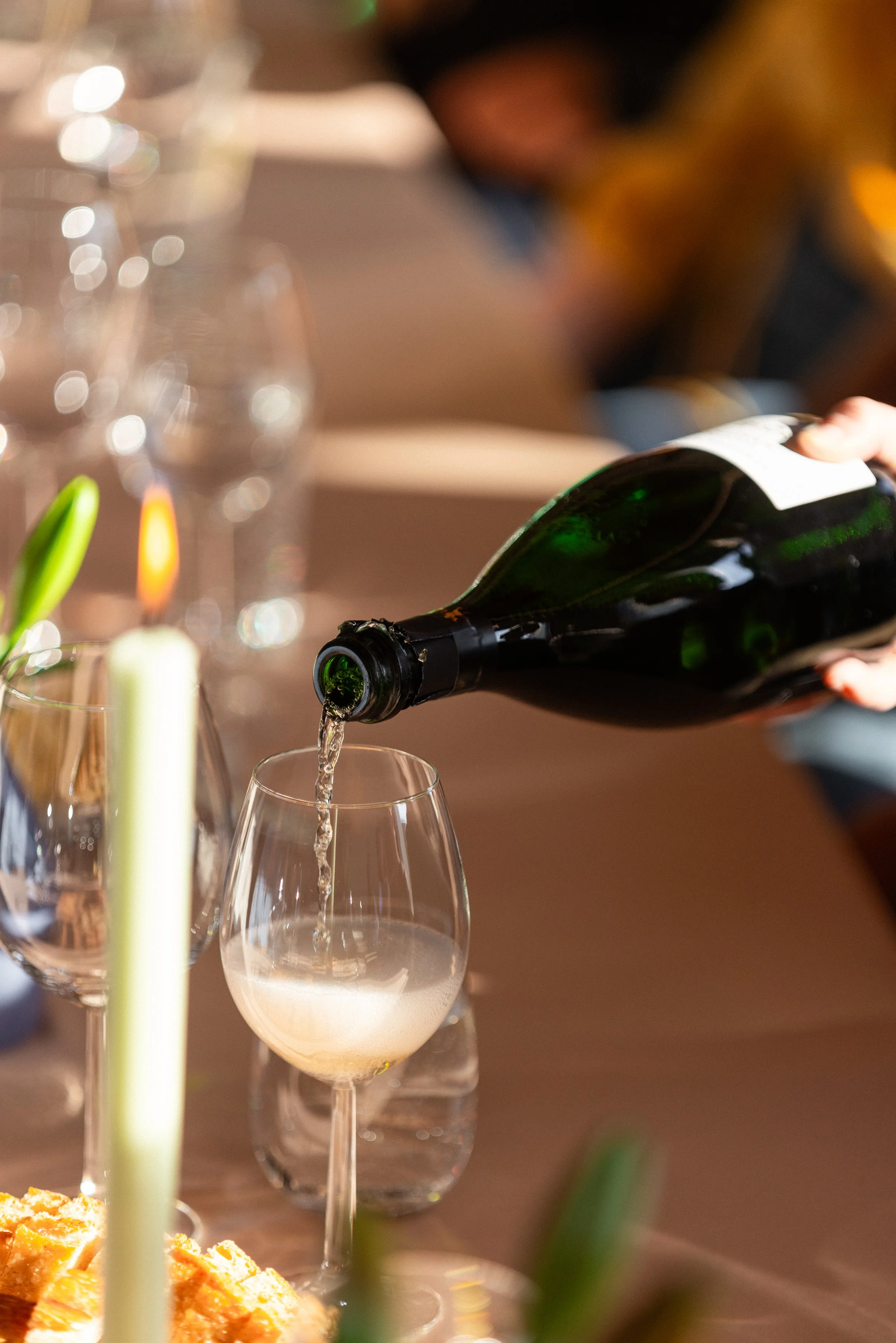 Person som heller sprudlende champagne i et glass ved et bord dekket med glass, blomster og mat.