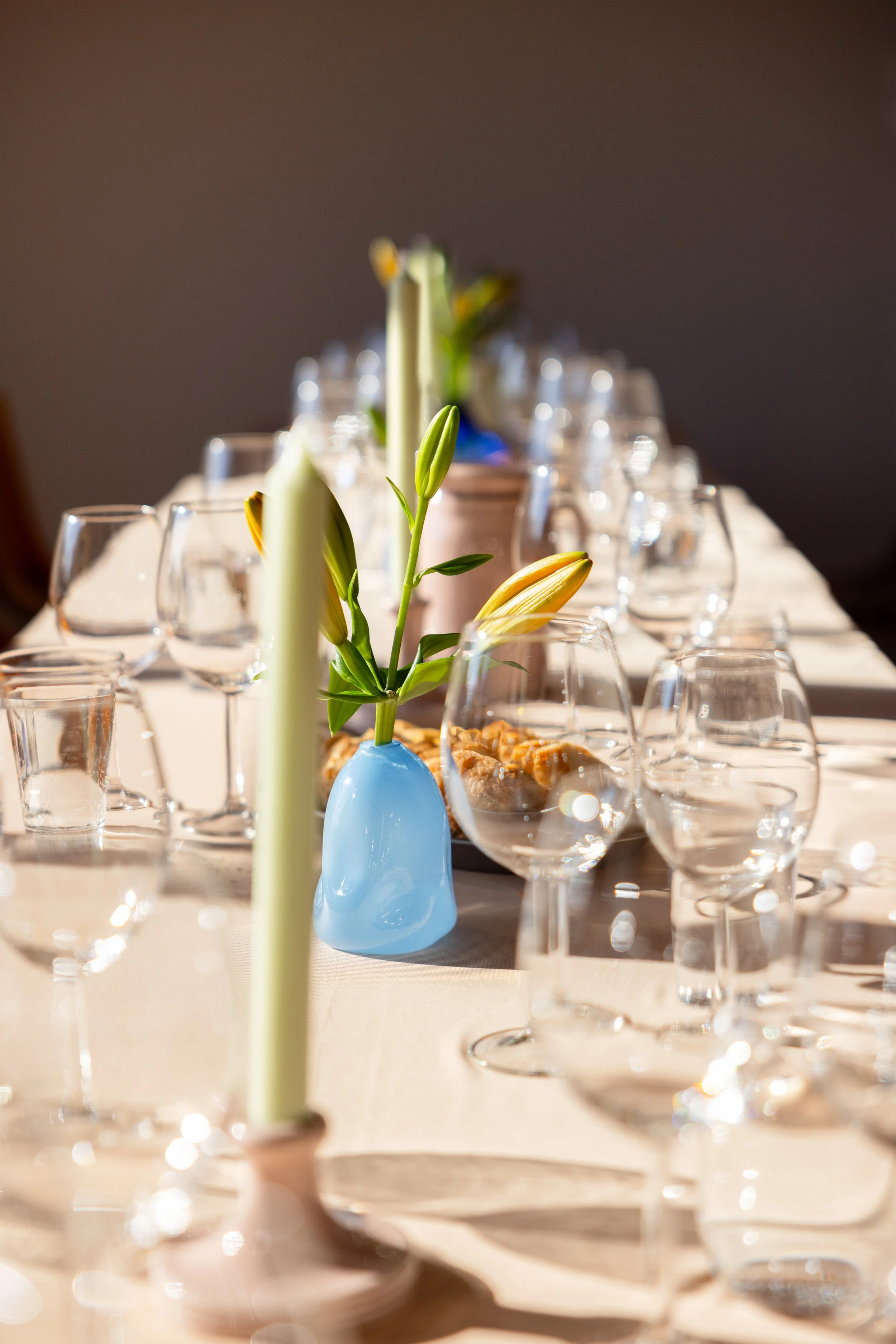 Elegant dekket bord med mange tomme vinglass, en blå vase med blomster, lys i lys rosa holder, og små tallerkener med snacks, i solfylt rom.