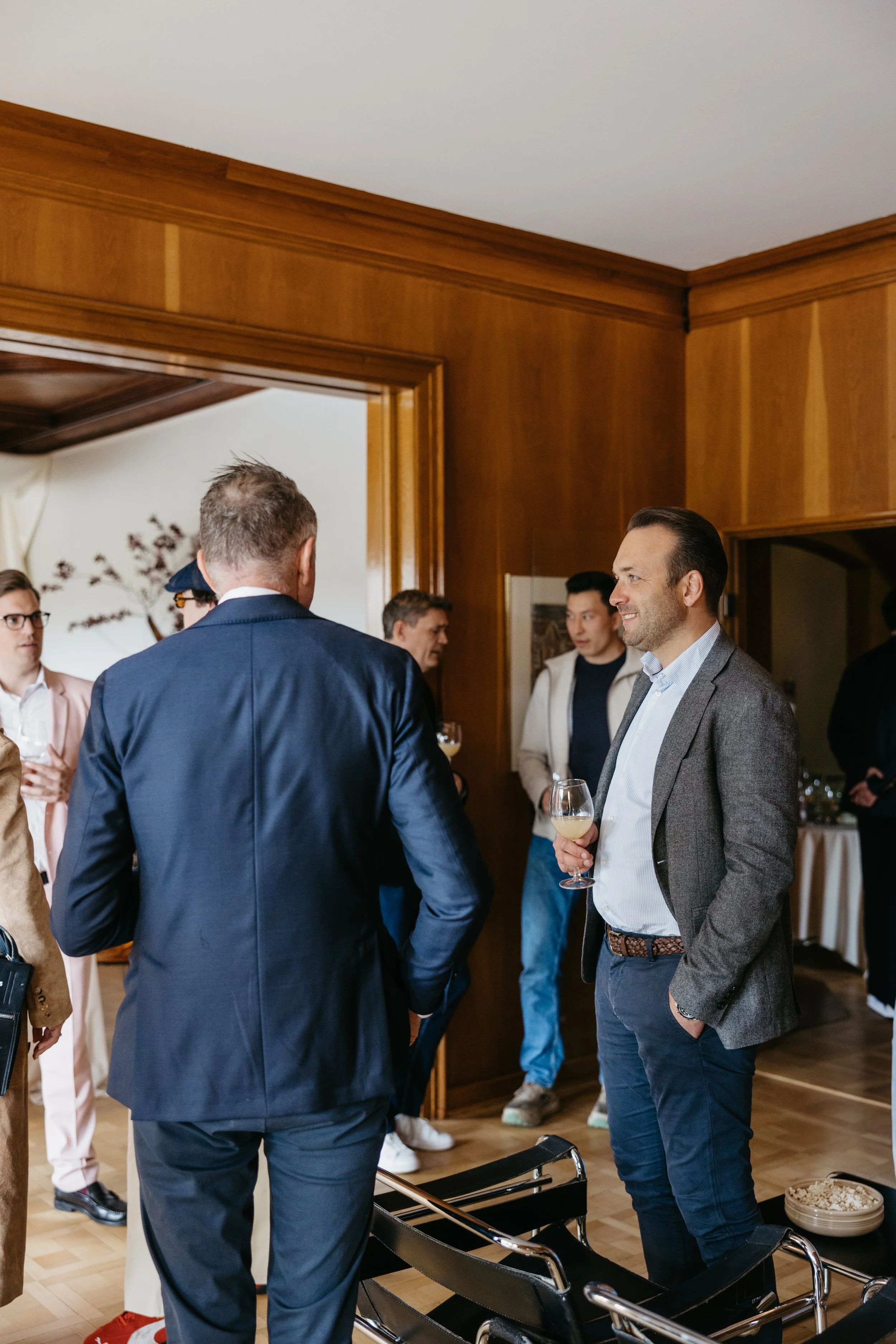 Mann med grå blazer som snakker med man i mørk blazer med glass vin i hånd under en sosial sammenkomst innendørs.