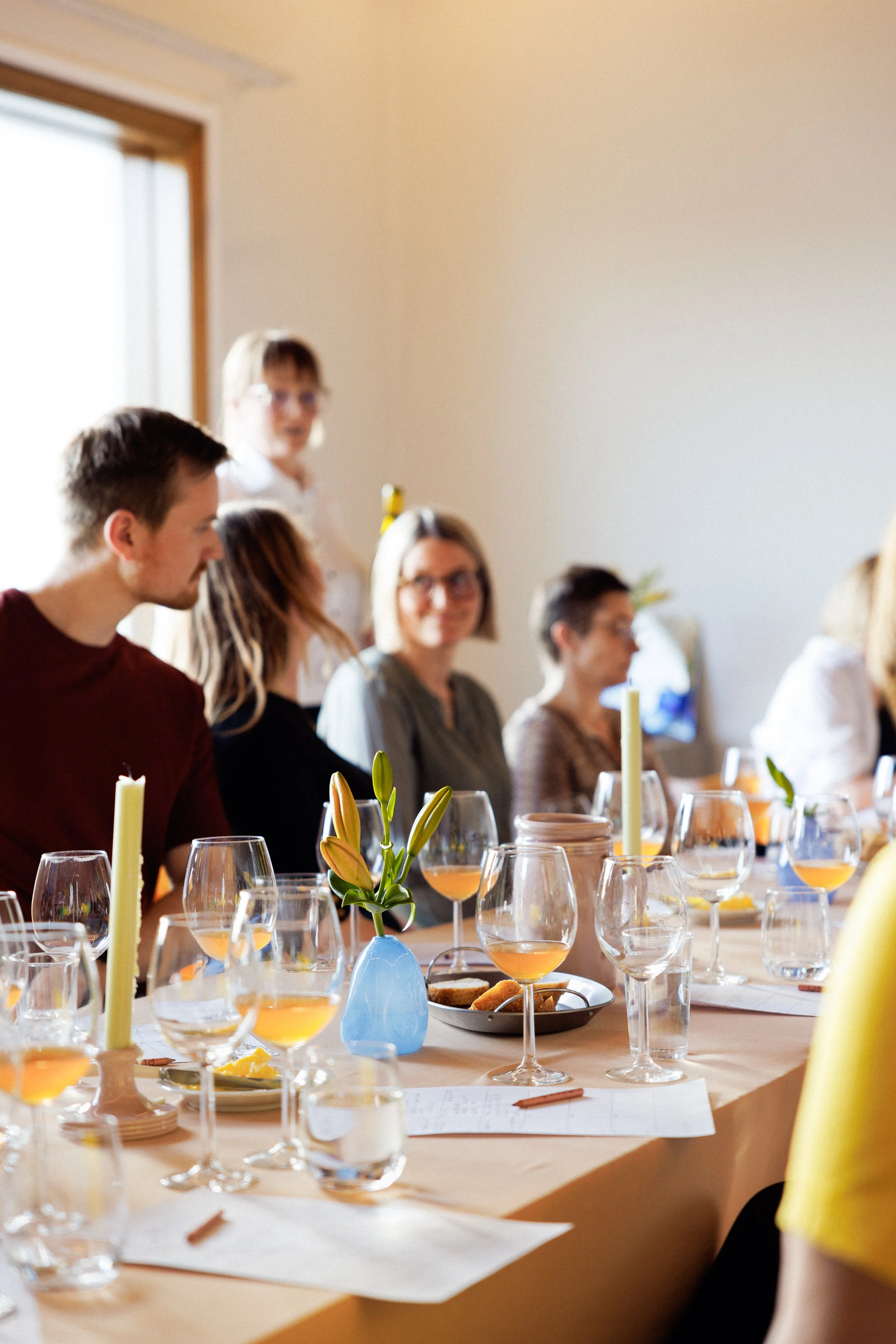 En gruppe mennesker sitter rundt et bord med blomster, stearinlys og glass med drikke, i et rom med lyse vegger og et vindu.
