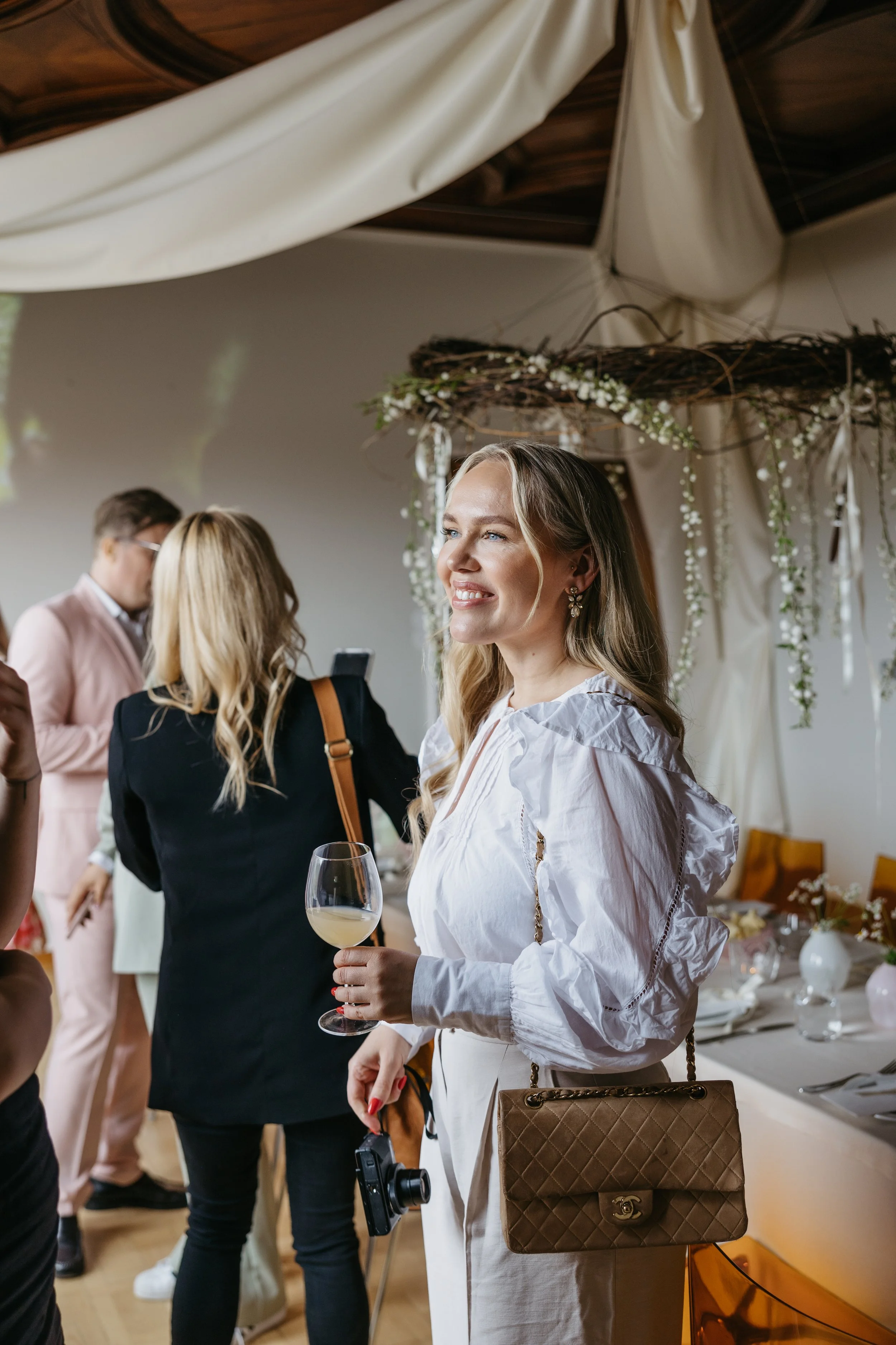 En smilende kvinne med langt blondt hår, iført en hvit bluse og gullfargede øredobber, holder et glass hvitvin og en kamera, og står i et festlig miljø med andre personer og blomsterdekorasjoner.