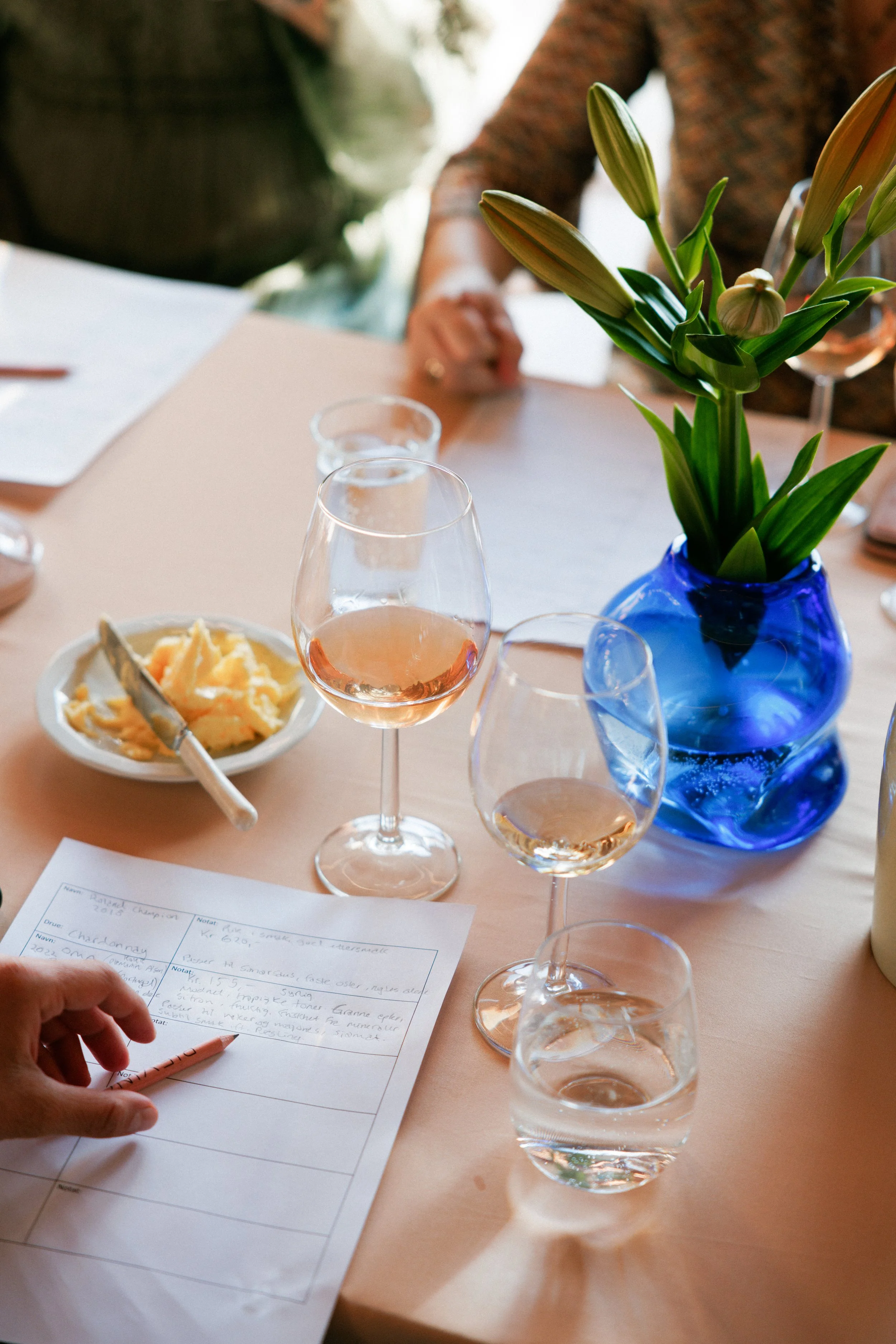 Et bord med to glass rosévin, en skål med smør og en vase med blå glass fylt med blomster, med mennesker i bakgrunnen.