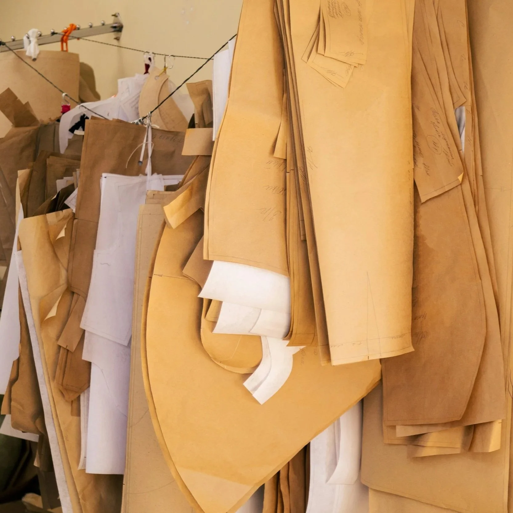 A large pile of pattern paper, mostly in beige and white, with some sketches and markings. Some papers are stacked, folded, or hanging over each other.