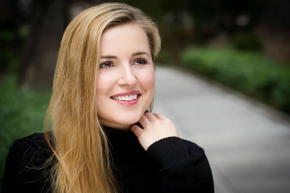 A smiling woman with long blonde hair, wearing a black top, outdoors with a blurred background of trees and a pathway.