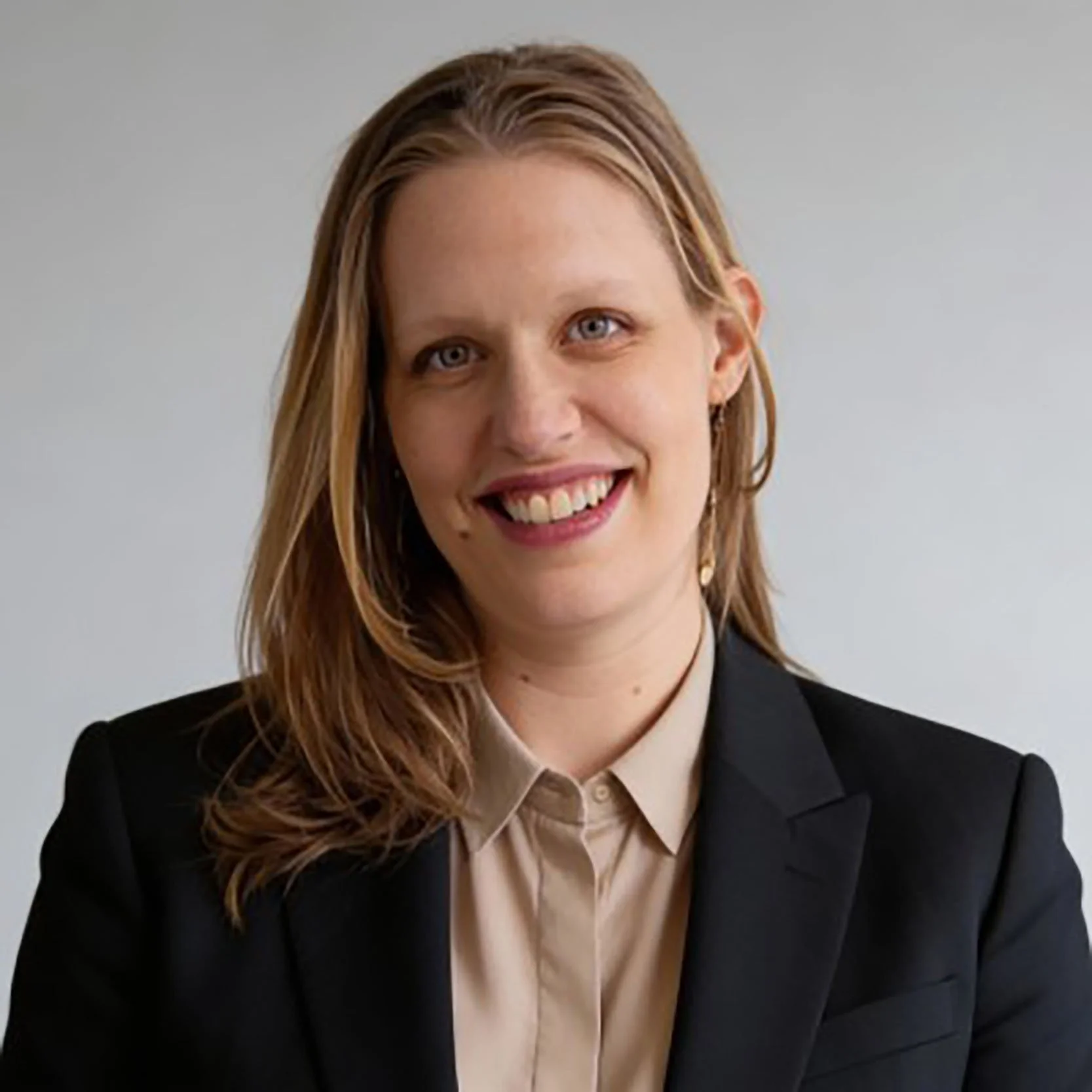 Headshot of a young woman with light brown hair, blue eyes, smiling, wearing a black blazer and beige shirt against a gray background.