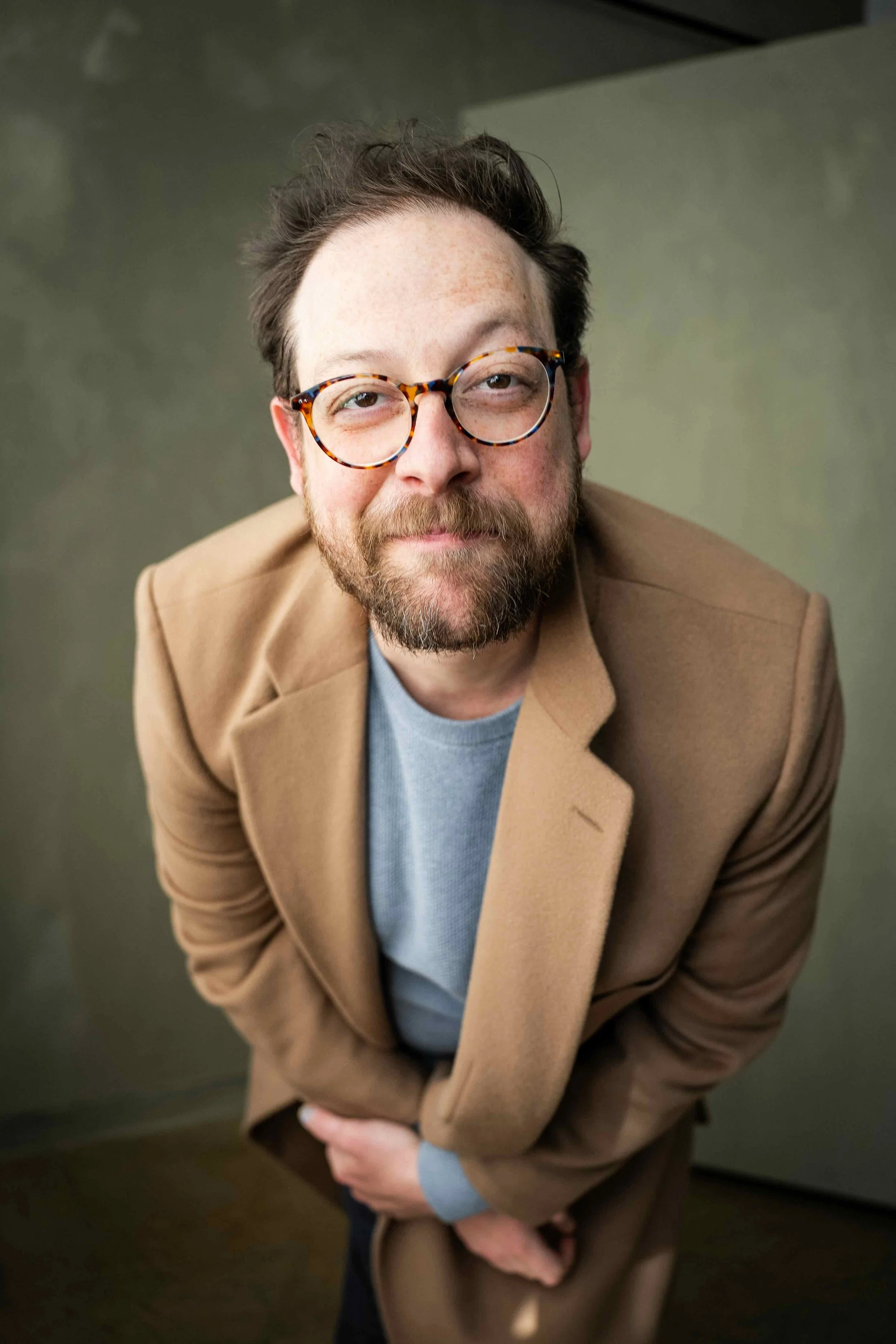A man with glasses, a beard, and curly hair wearing a brown coat and a light gray shirt, looking up at the camera with a slight smile.