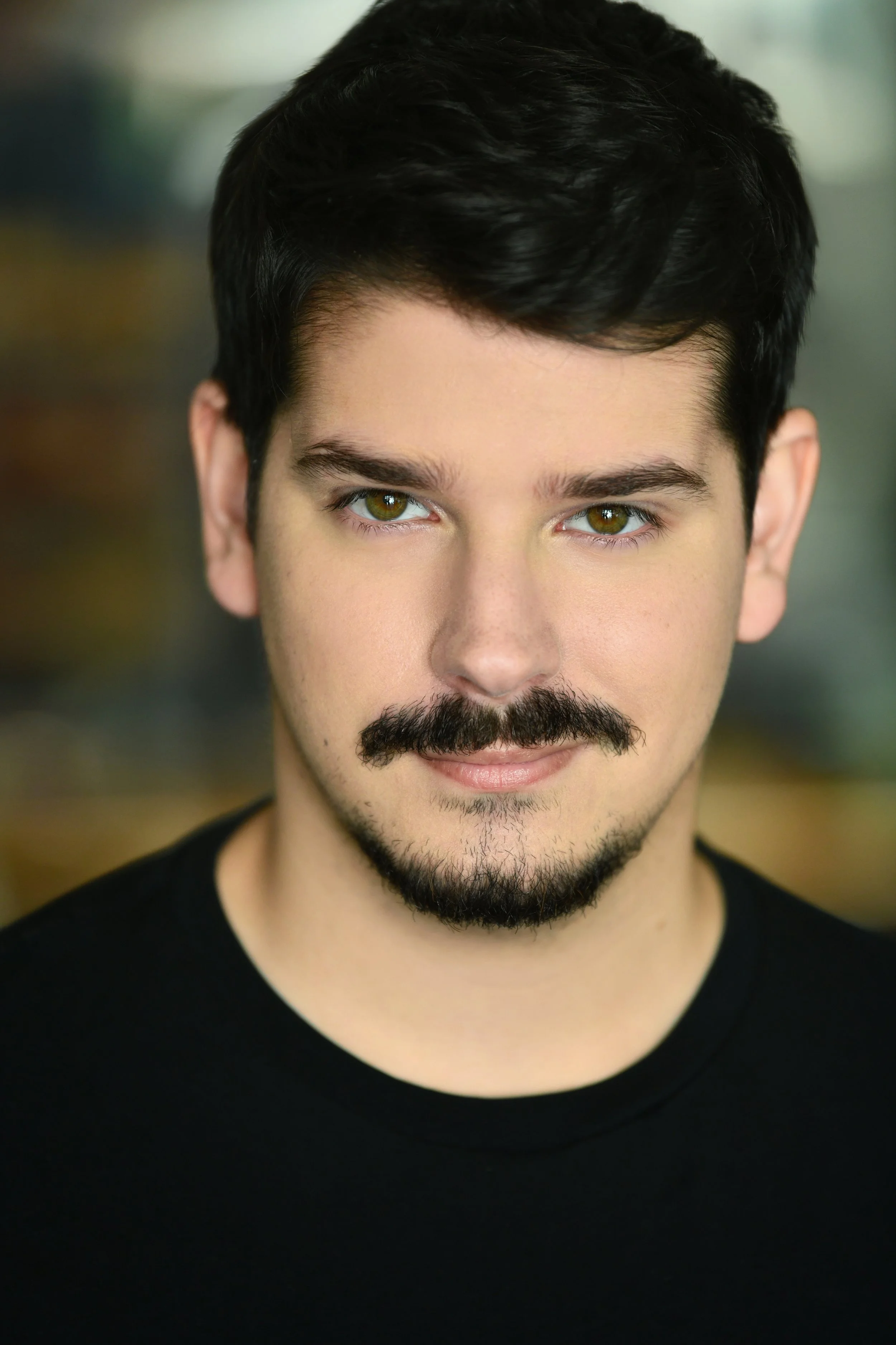 Close-up of a young man with dark hair, hazel eyes, and a trimmed beard, wearing a black shirt, smiling slightly at the camera.