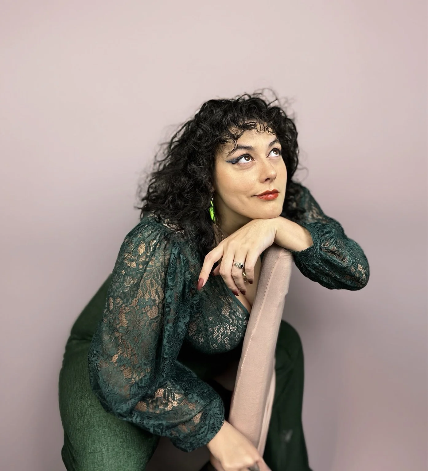 A woman with curly black hair and makeup, wearing a green lace top with long sleeves, is leaning on a beige chair with her chin resting on her hand against a pink wall background.