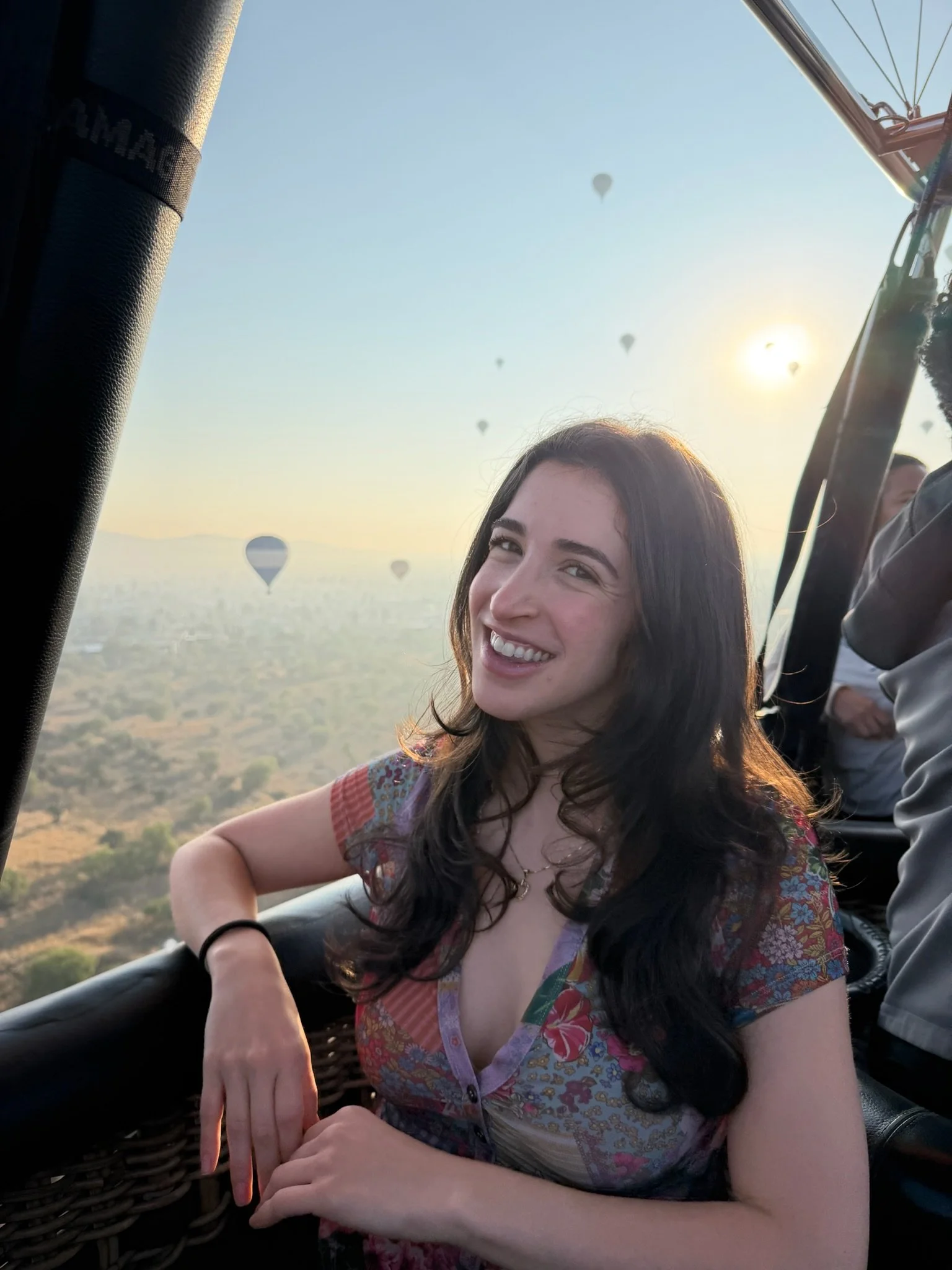 A woman with dark hair and a patterned dress sitting in a hot air balloon basket, smiling at the camera with a landscape and other hot air balloons in the background as the sun sets.