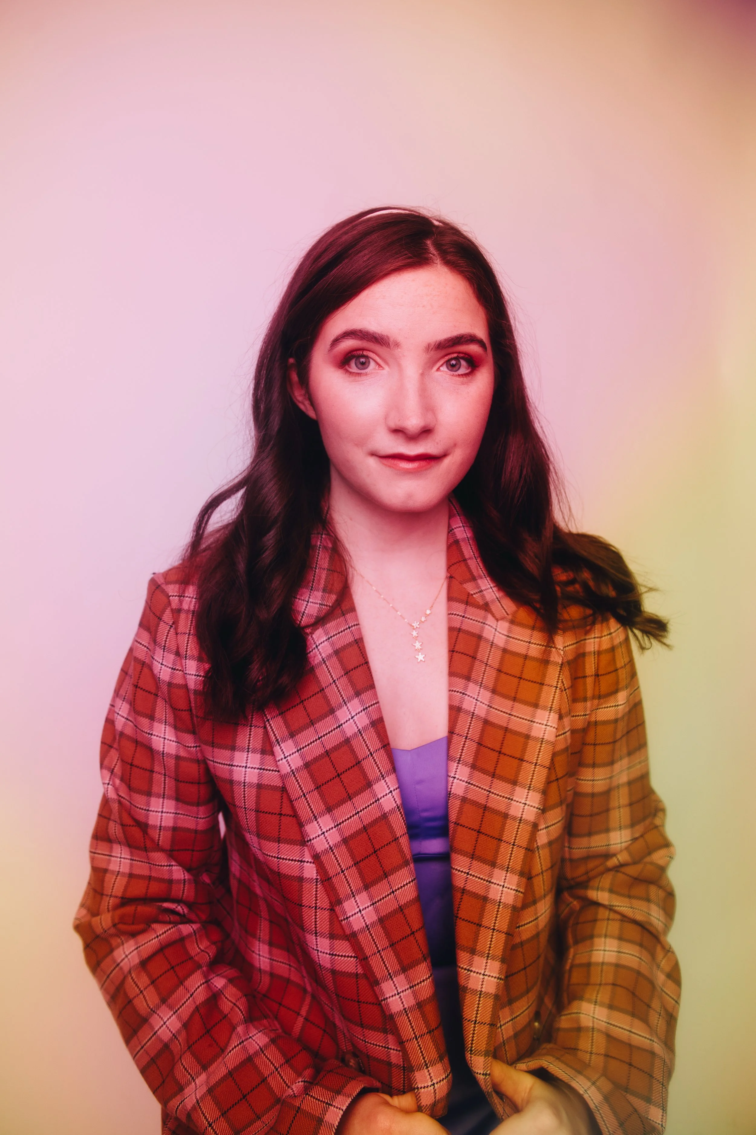 A young woman with long dark hair wearing a plaid orange and brown blazer against a multicolored background.