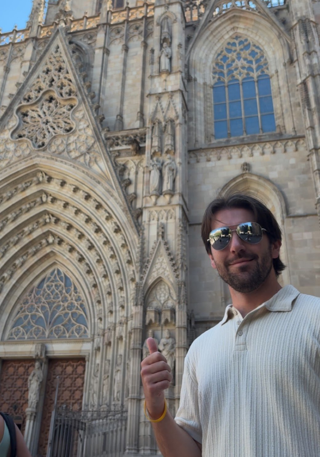 A man with sunglasses and a light-colored striped shirt giving a thumbs up in front of a Gothic cathedral with ornate stone carvings and large stained glass windows.