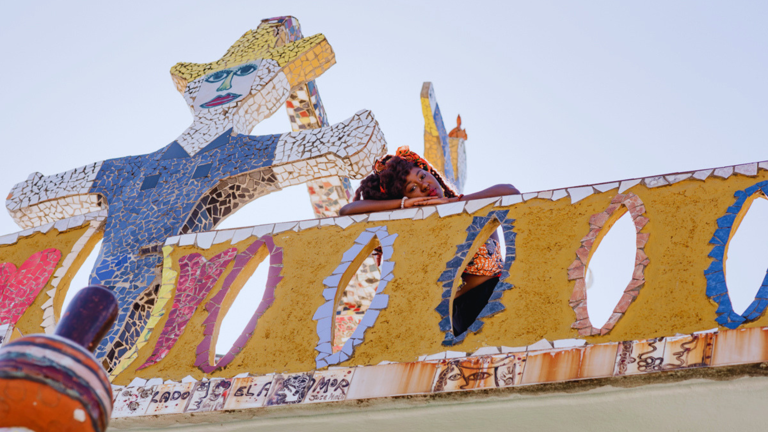 A woman with curly hair and colorful clothing lying on a decorated, mosaic-covered sculpture with a face and arm, set against a clear sky.