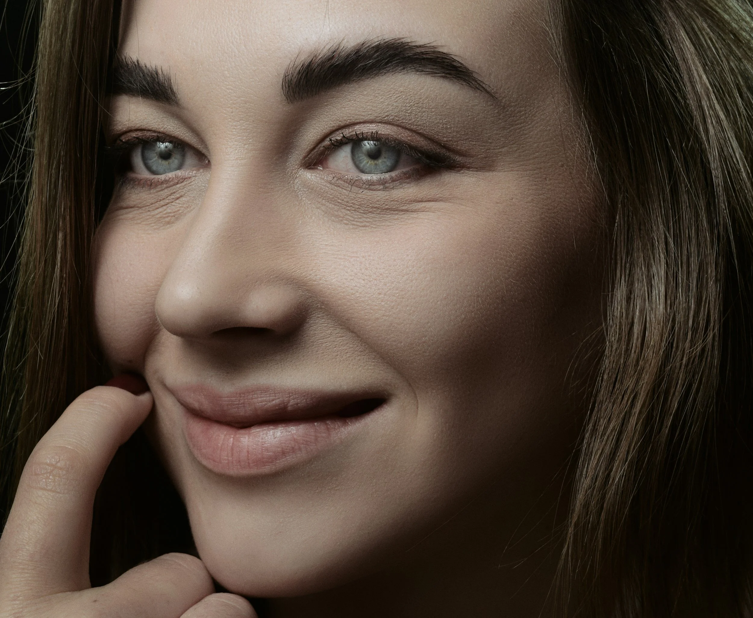 Close-up of a woman's face, showing her eyes, eyebrows, and part of her nose and lips. She is touching her lips with her fingers and has long brown hair.