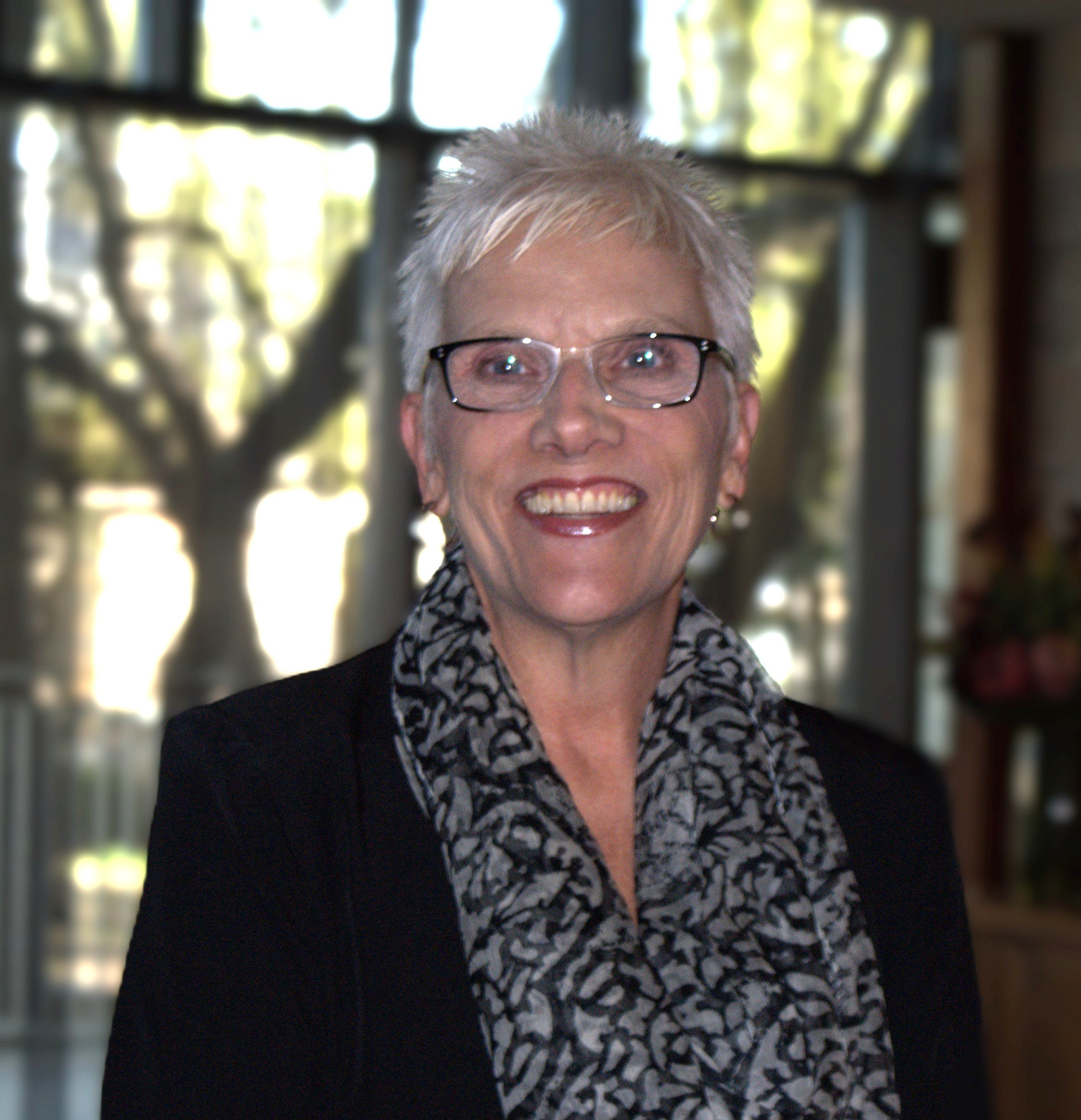 A woman with short white hair, glasses, and gold hoop earrings is smiling and wearing a beige blazer. She is standing in front of a glass window with trees visible outside.
