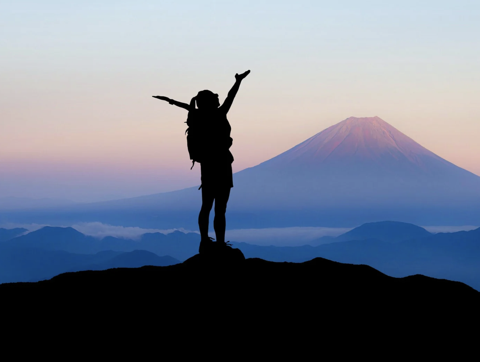 Silhouette of a person with a backpack standing on a mountain summit, arms raised, with Mount Fuji and a colorful sky in the background at sunrise or sunset.